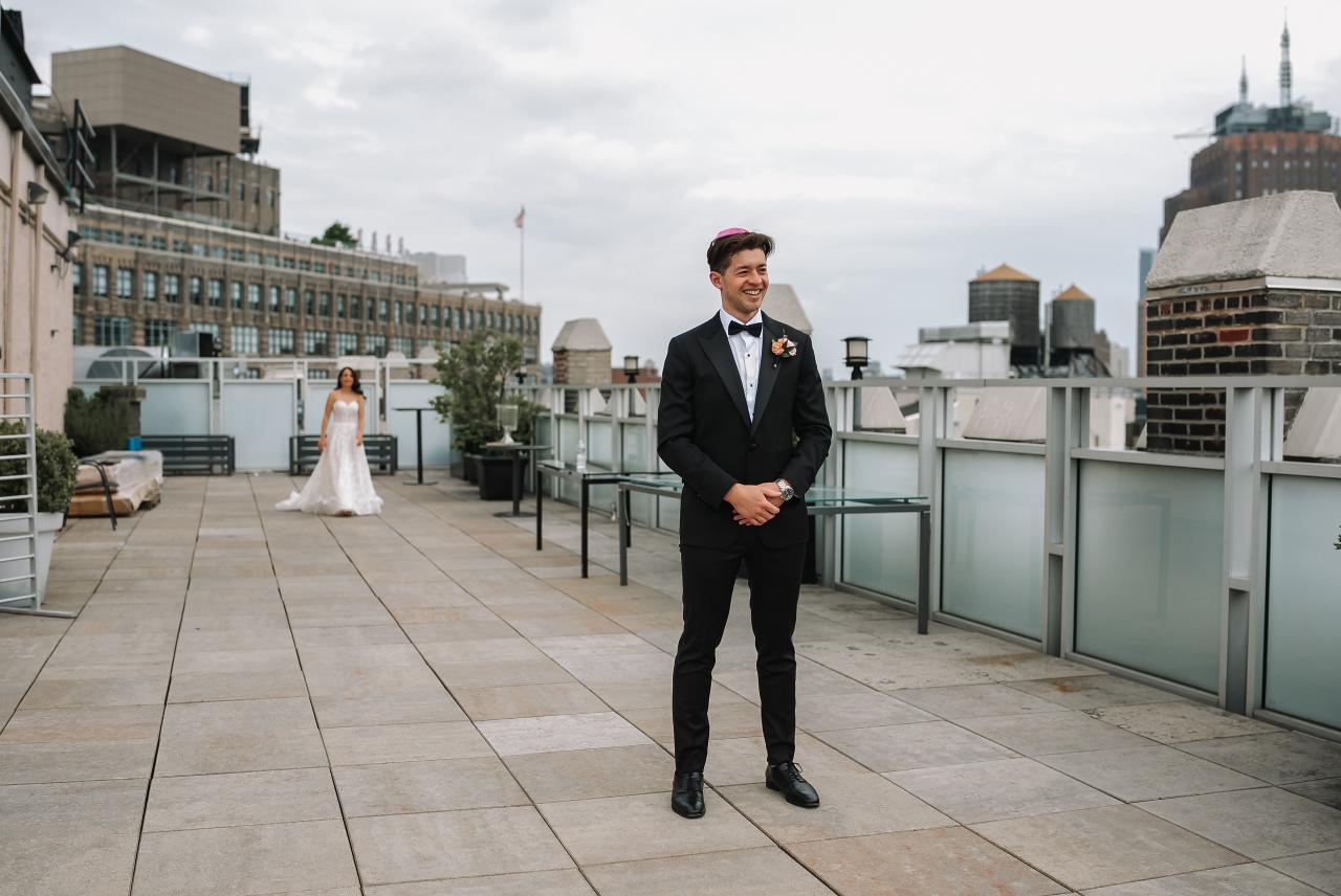 Groom in a black suit smiling on an outdoor terrace walkway with the bride in a white wedding dress in the background. Wedding photography at Tribeca Rooftop + 360°, New York, NY.