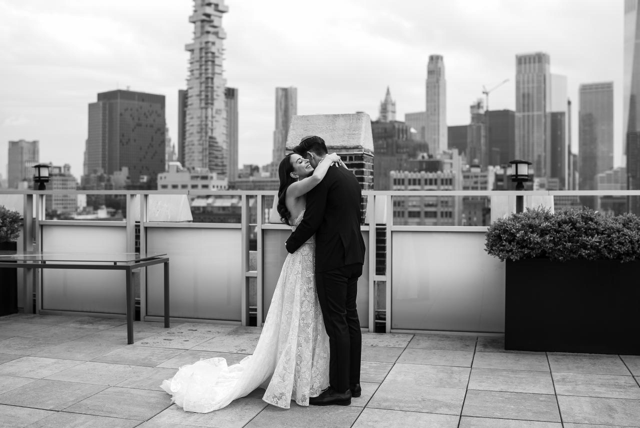 Black-and-white rooftop wedding portrait of a couple embracing with the city skyline behind them; bride’s lace train trails across the stone floor. Wedding photography at Tribeca Rooftop + 360°, New York, NY.