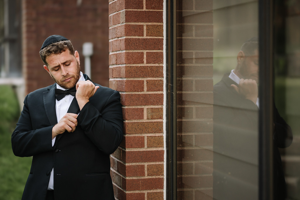 A groom in a black tuxedo adjusts his cuff and collar near a brick wall and window before the ceremony.