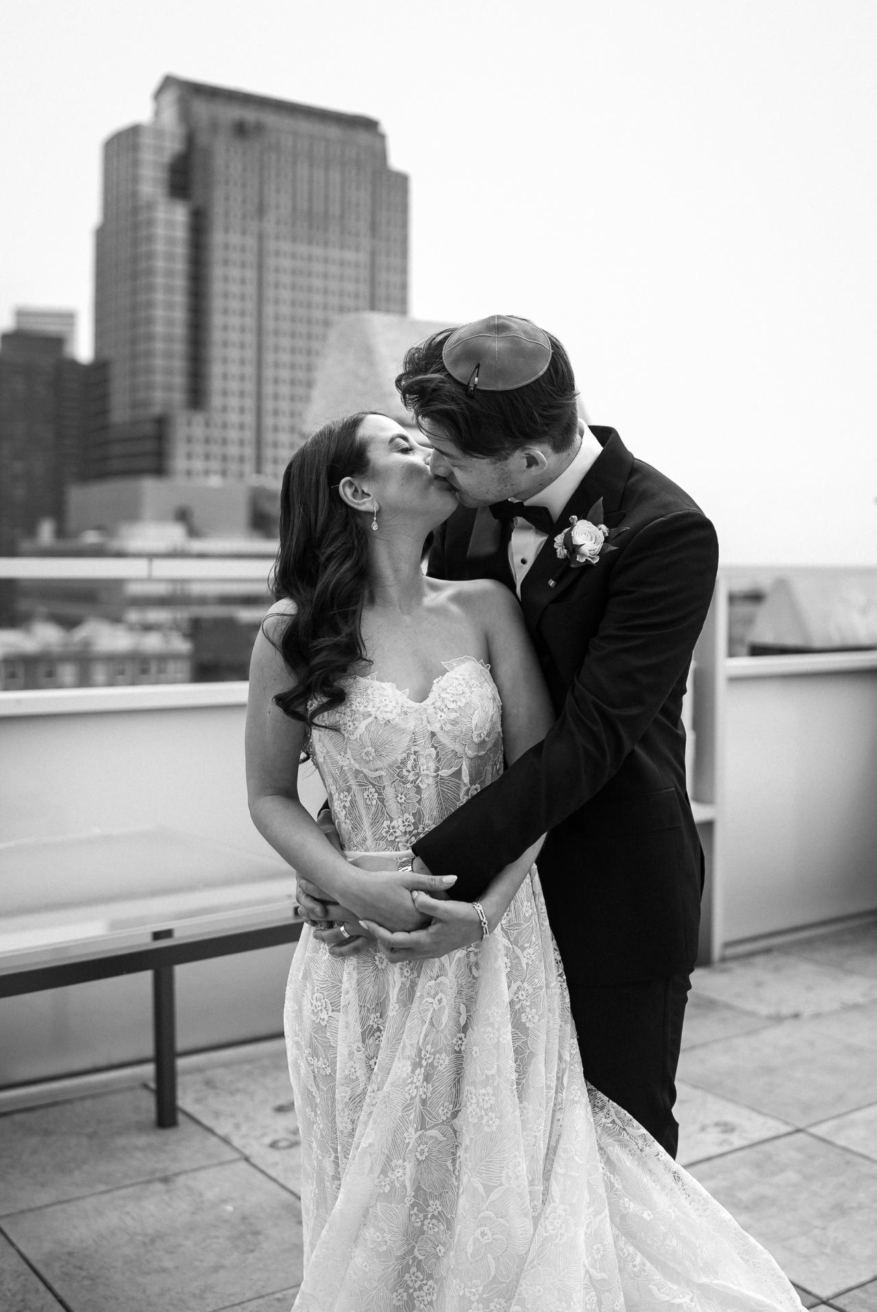 Black-and-white photo of a bride and groom kissing while embracing on a rooftop terrace, with a city skyline in the background. Wedding photography at Tribeca Rooftop + 360°, New York, NY.