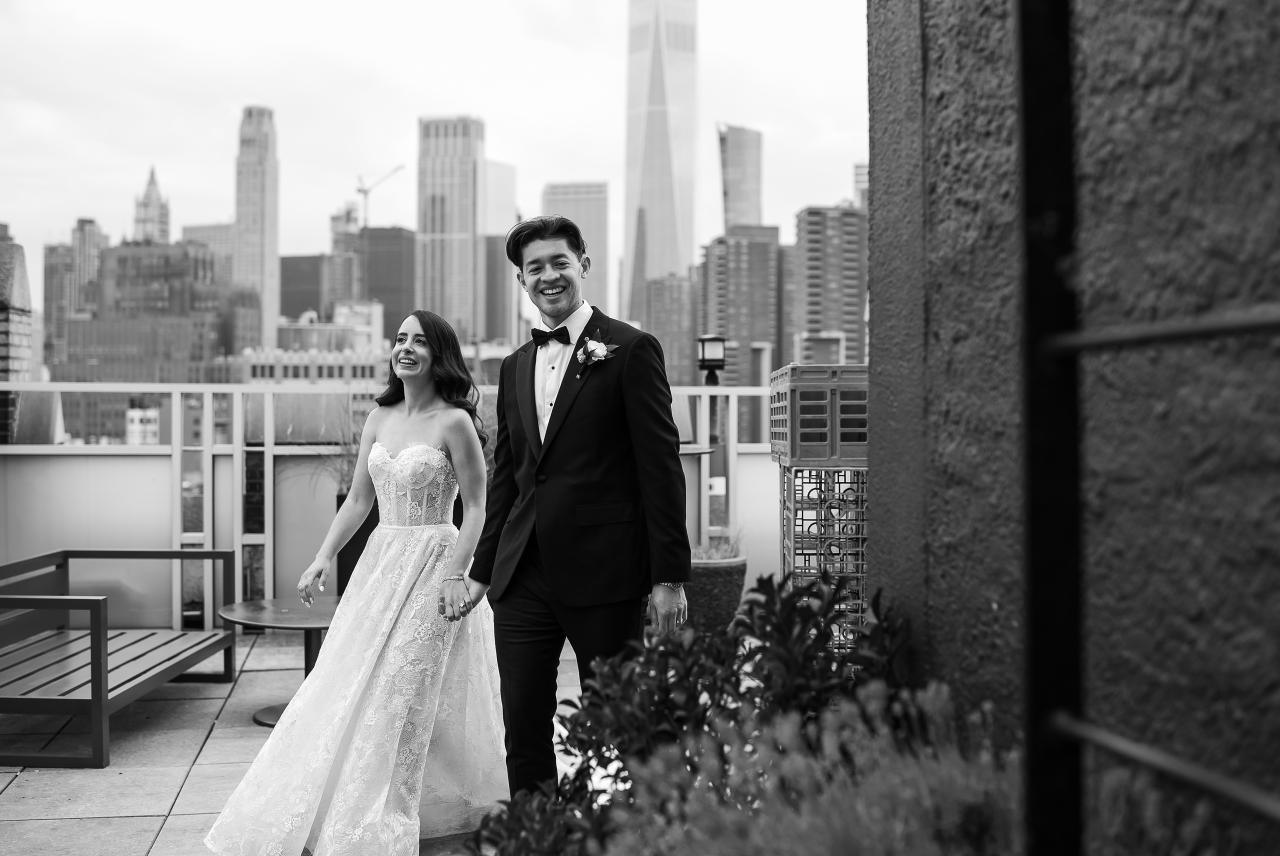 Black-and-white photo of a bride and groom walking on a rooftop balcony, smiling with a city skyline behind them. Wedding photography at Tribeca Rooftop + 360°, New York, NY.