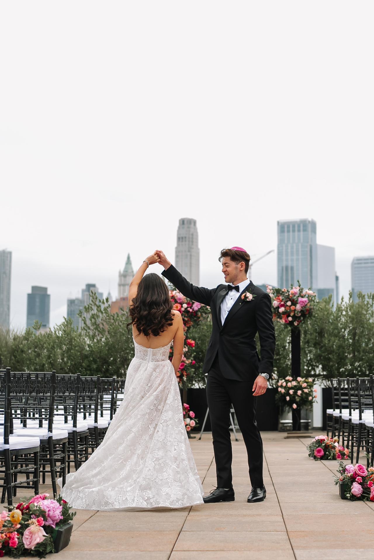 A joyful wedding couple celebrating at an outdoor ceremony aisle, with skyline buildings in the background and floral arrangements beside the chairs. Wedding photography at Tribeca Rooftop + 360°, New York, NY.
