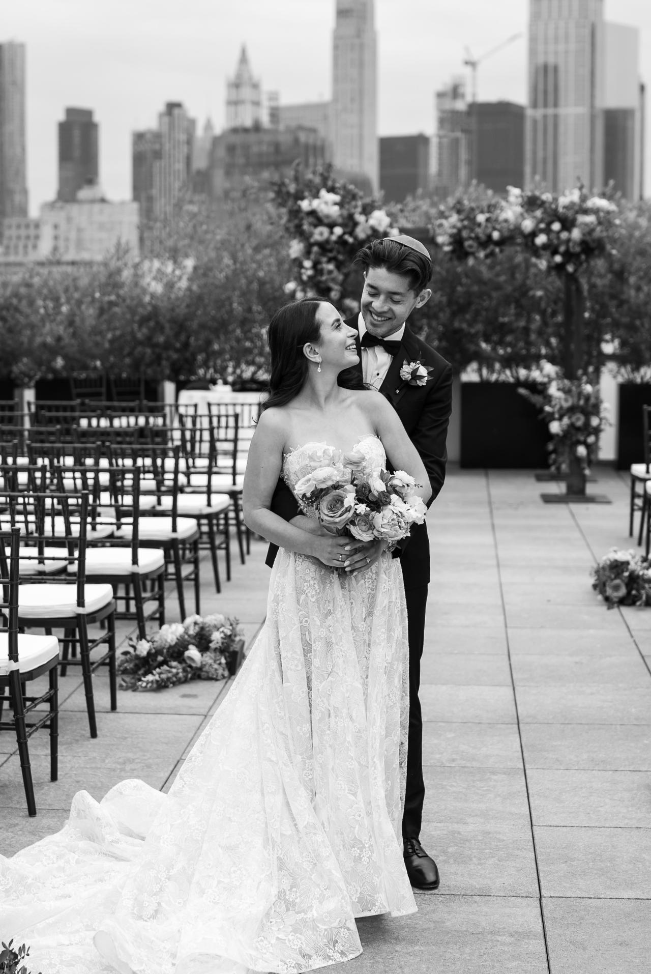 Black-and-white outdoor wedding portrait of a bride and groom standing close together and smiling, holding a bouquet, with flowers and a city skyline in the background. Wedding photography at Tribeca Rooftop + 360°, New York, NY.