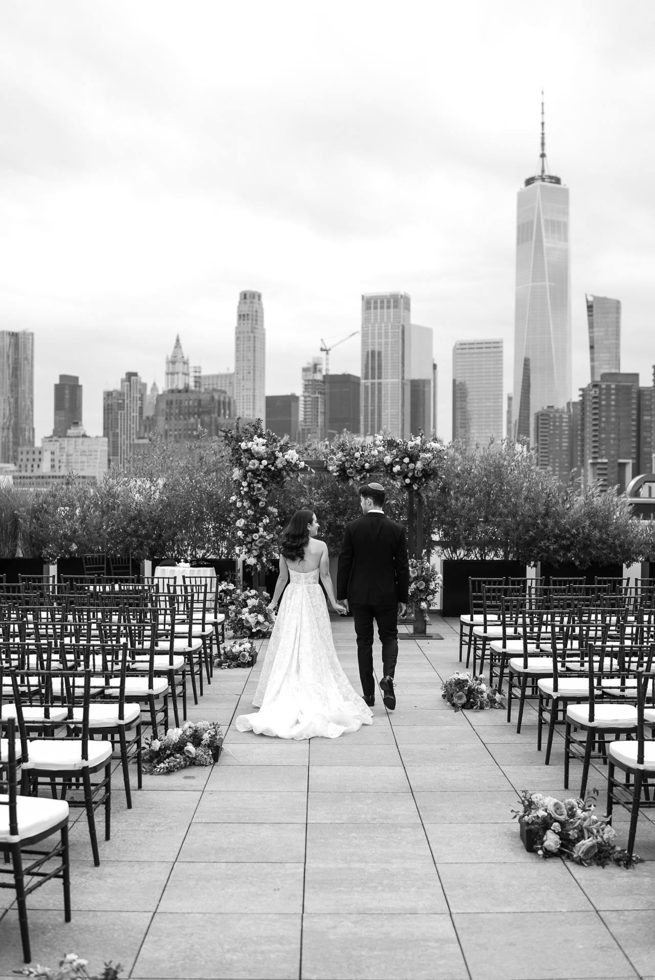 Black-and-white wedding ceremony scene with a bride and groom walking hand in hand along an aisle between chairs, with flower arrangements and a city skyline in the background. Wedding photography at Tribeca Rooftop + 360°, New York, NY.
