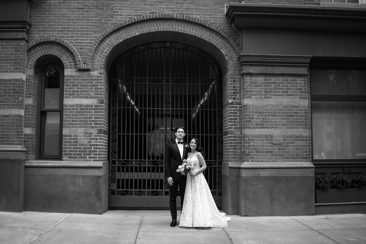 Black-and-white wedding portrait of a bride and groom standing together in front of a stone building with an arched iron gate. Wedding photography at Tribeca Rooftop + 360°, New York, NY.