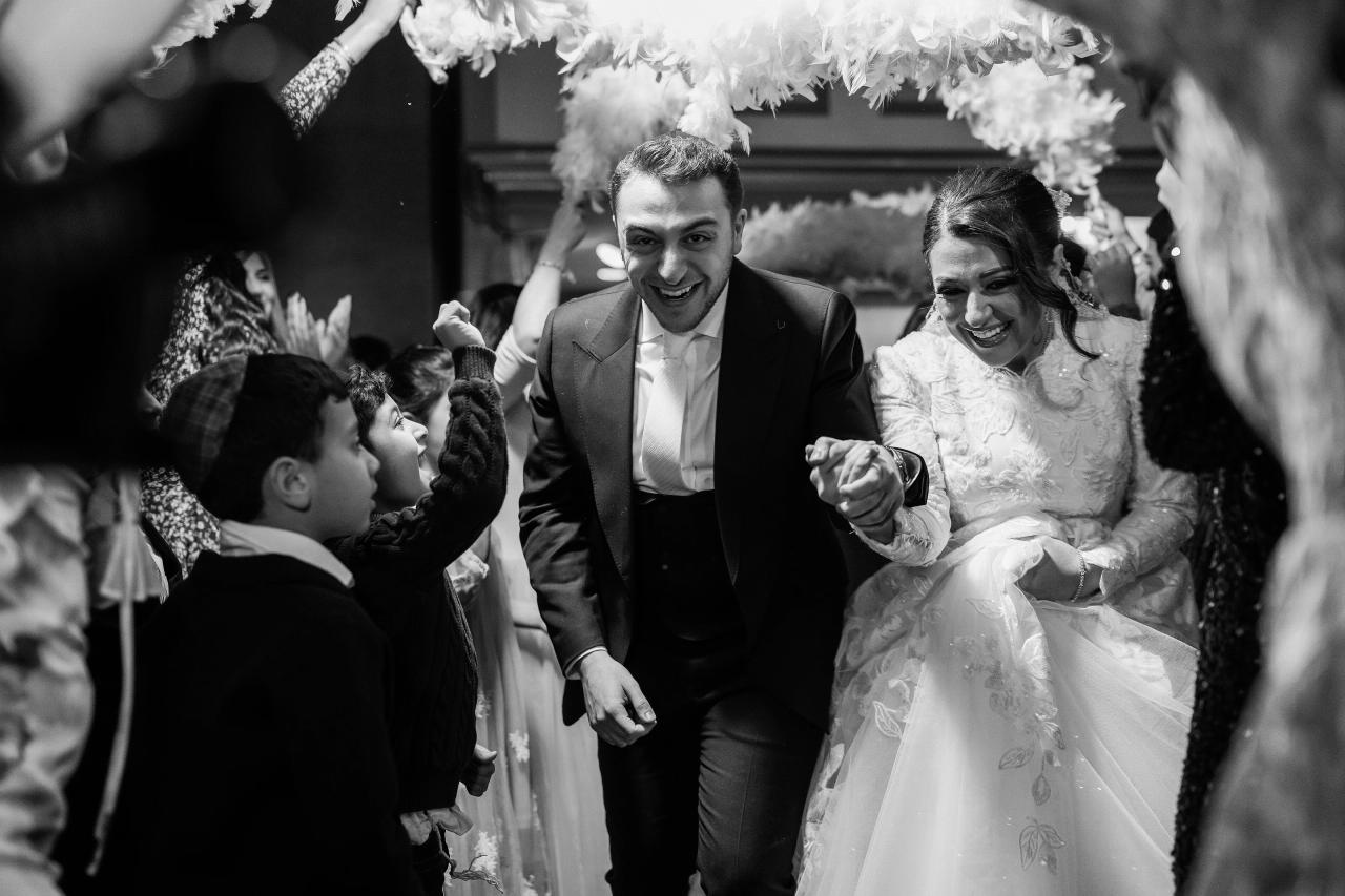 Black-and-white candid of the groom and bride smiling under the chuppah while a child in the foreground raises his arm and reaches toward them at a wedding celebration.