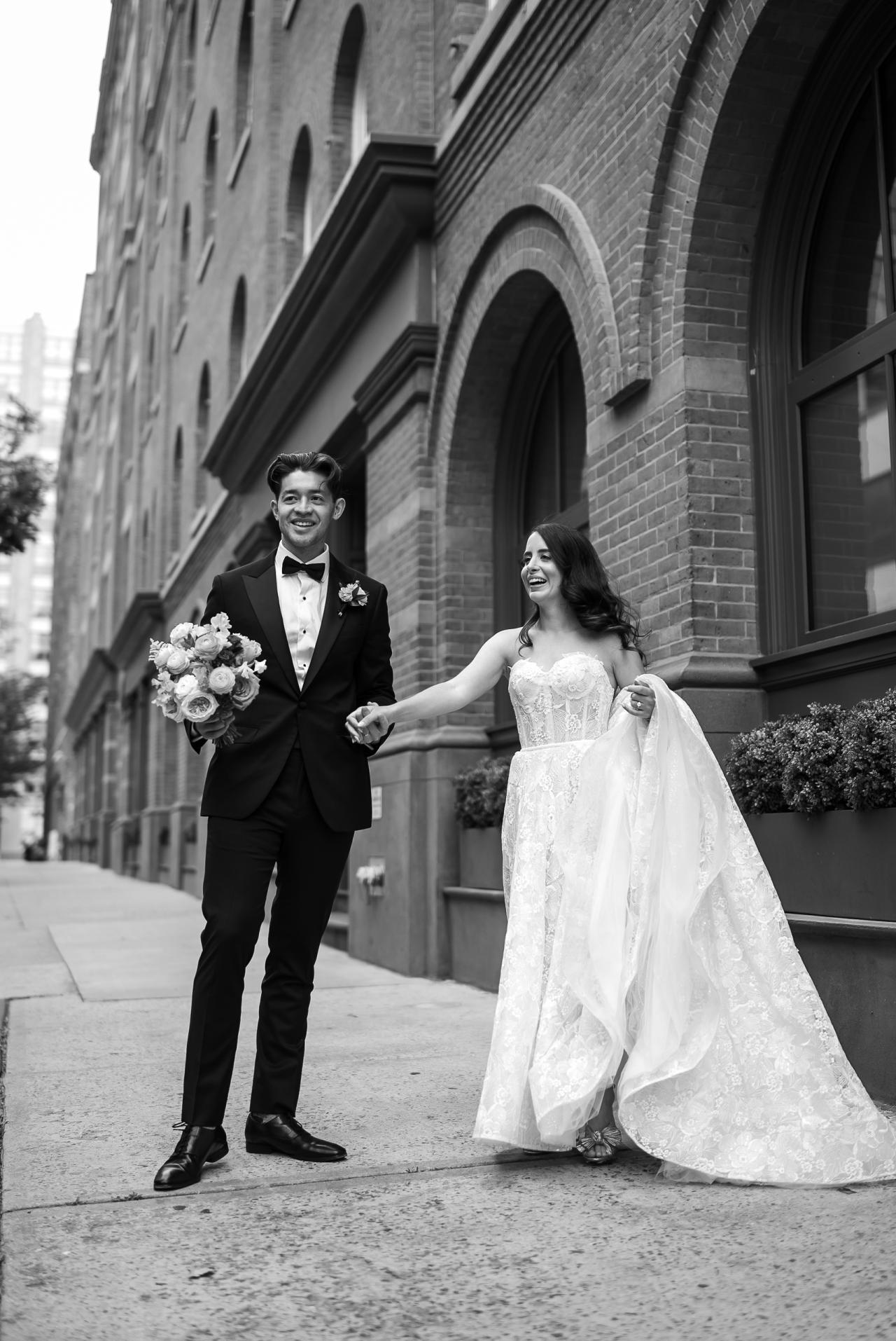 Black-and-white wedding portrait of a smiling couple walking hand-in-hand beside arched brick architecture, bride in a lace gown holding her train, groom holding a bouquet. Wedding photography at Tribeca Rooftop + 360°, New York, NY.