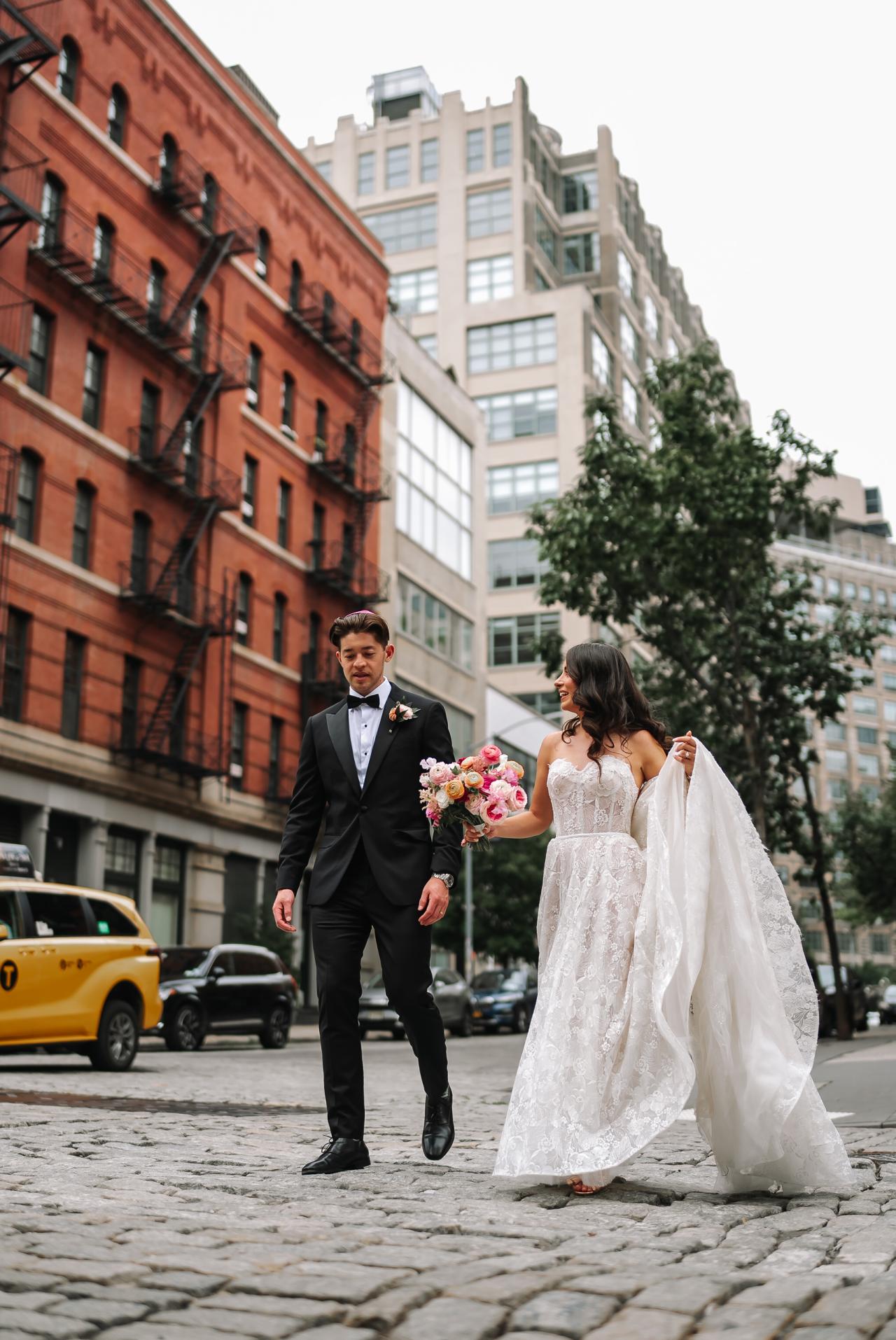 Bride and groom walking together outdoors on a cobblestone street, bride holding a bouquet, framed by tall city buildings and greenery. Wedding photography at Tribeca Rooftop + 360°, New York, NY.