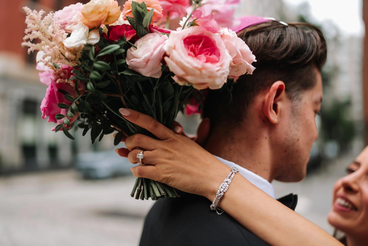 Bride and groom sharing a tender embrace outdoors while the bride holds a bouquet of pink and peach flowers in the foreground. Wedding photography at Tribeca Rooftop + 360°, New York, NY.