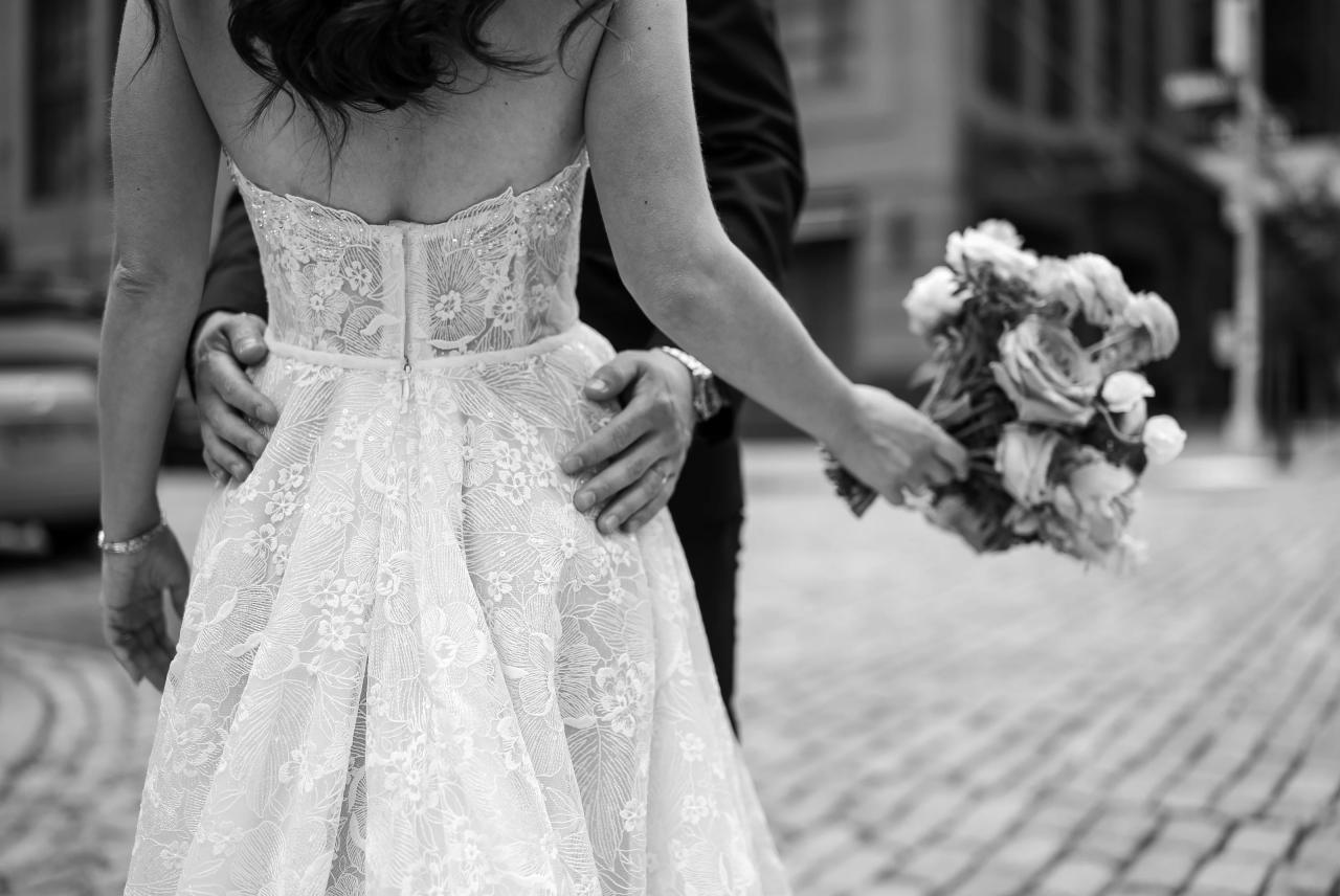 Black-and-white close-up of a bride’s hands holding her wedding dress at the waist while her groom stands beside her, with a bouquet visible in the background. Wedding photography at Tribeca Rooftop + 360°, New York, NY.