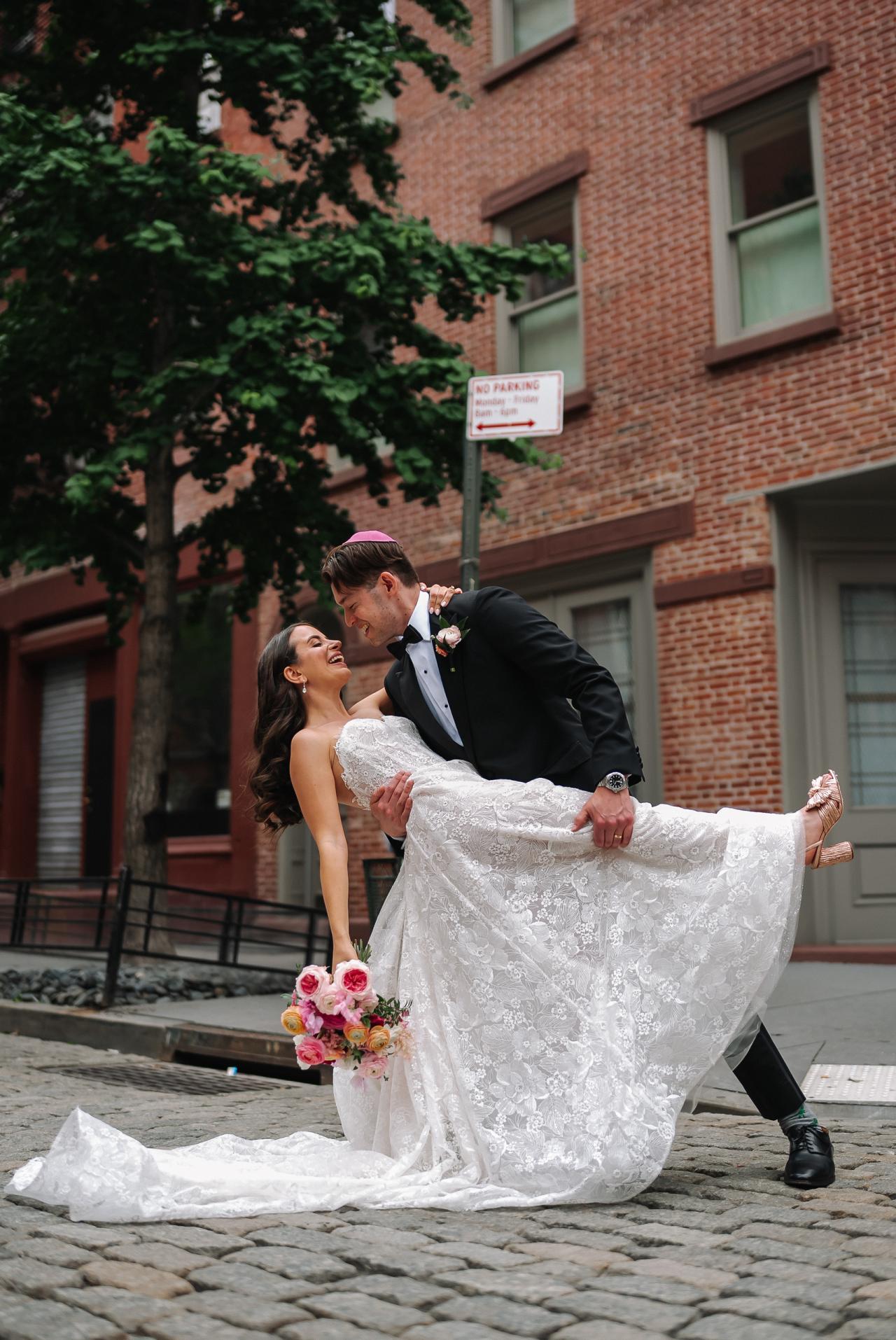 Bride and groom laughing while spinning outdoors on a cobblestone walkway in front of brick buildings and greenery; bride holds her bouquet as her dress fans outward. Wedding photography at Tribeca Rooftop + 360°, New York, NY.