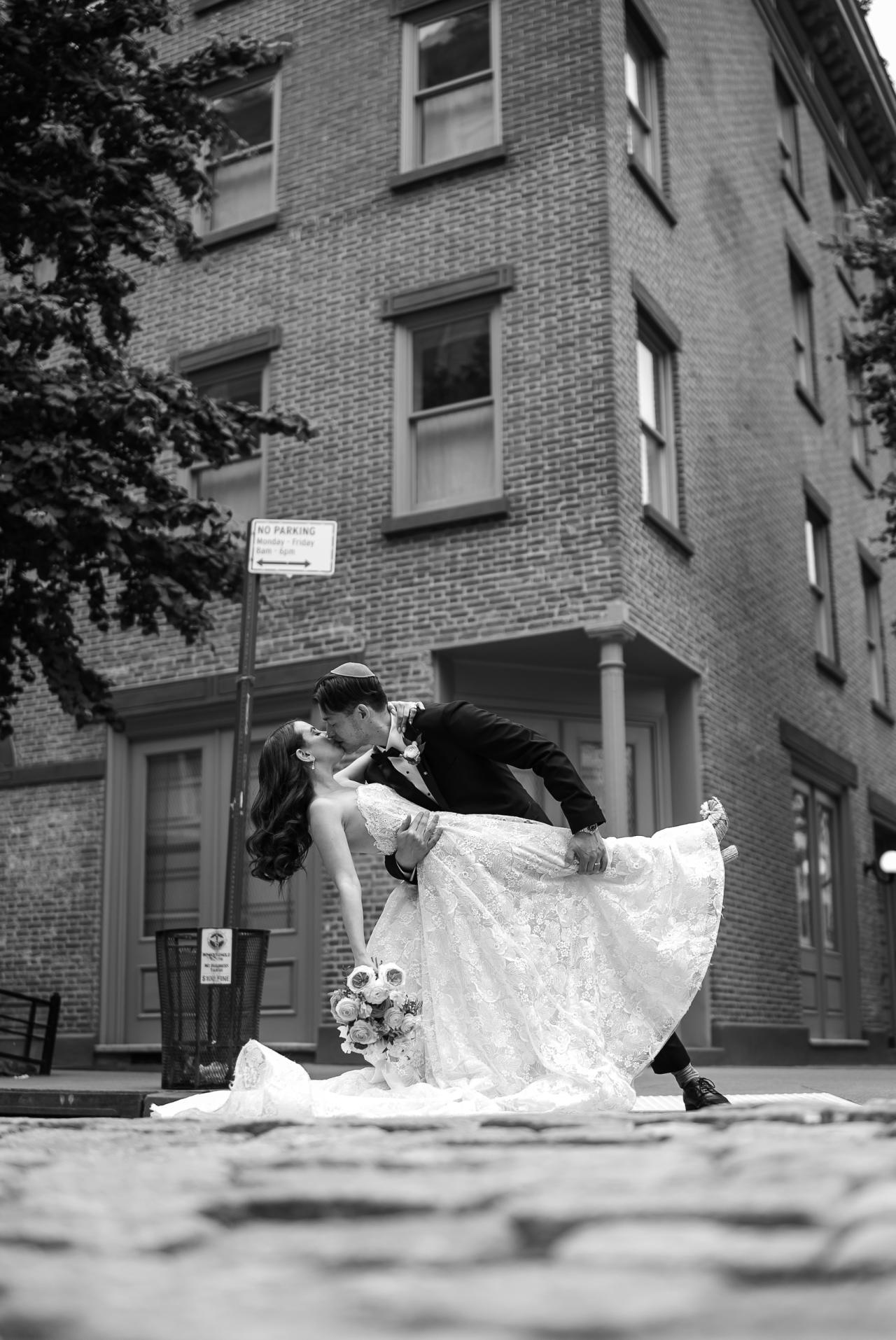 Black-and-white outdoor portrait of a bride and groom leaning into a kiss-like moment, bride’s flowing dress lifted as they pose in front of a brick building. Wedding photography at Tribeca Rooftop + 360°, New York, NY.