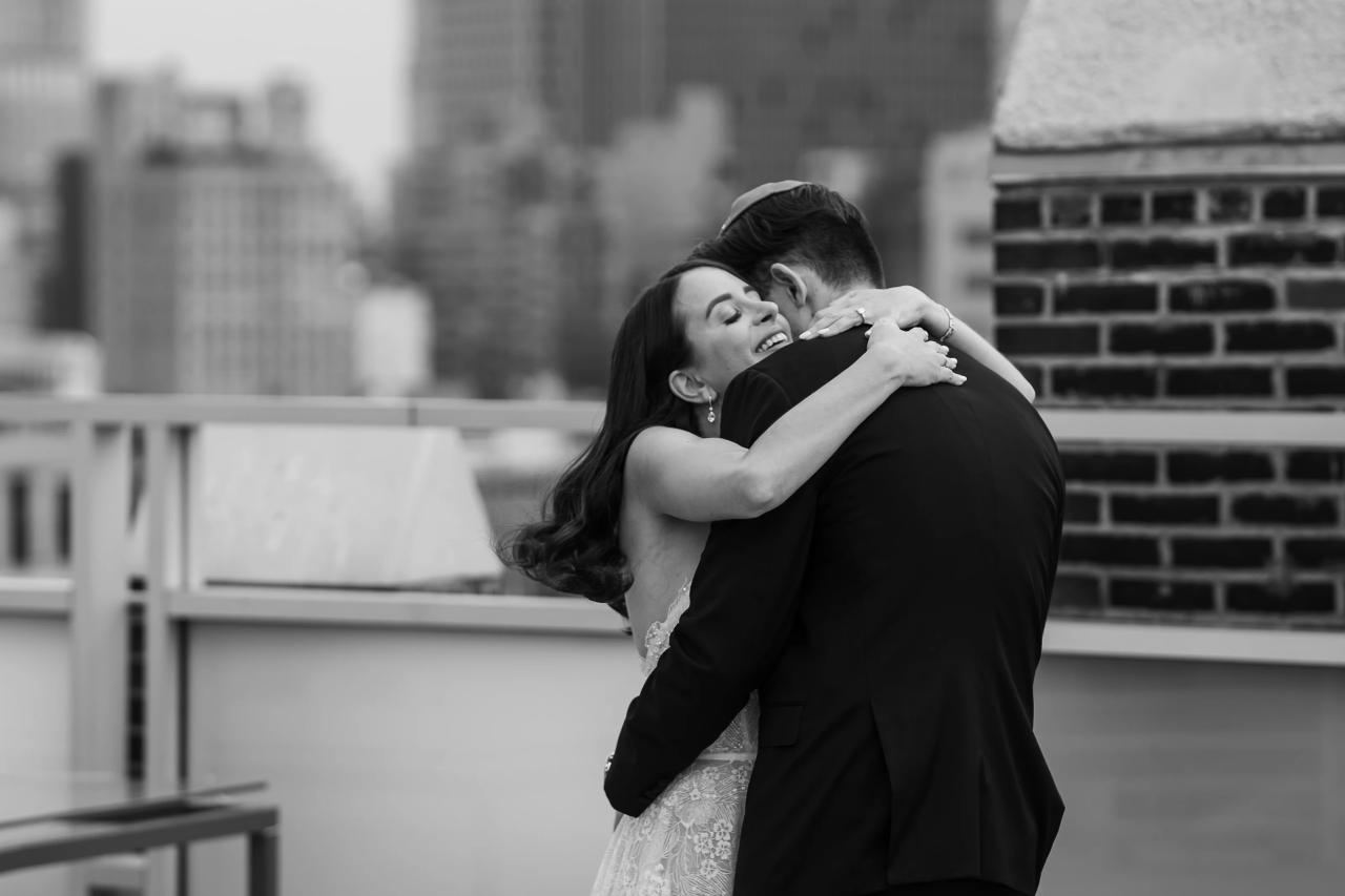 Black-and-white wedding portrait of a couple in a close embrace, bride laughing into the groom’s shoulder with modern brick and light architecture behind them. Wedding photography at Tribeca Rooftop + 360°, New York, NY.
