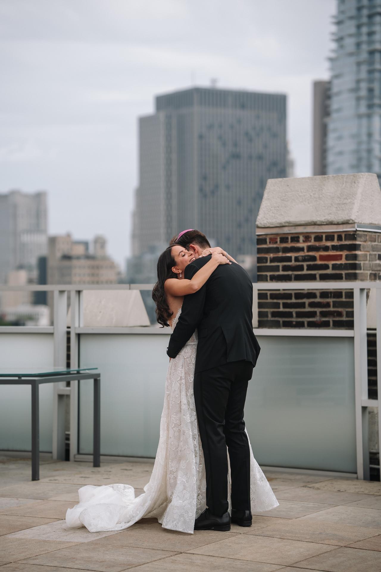 Bride and groom sharing an intimate embrace on a rooftop, bride’s gown flowing along the stone floor, city buildings softly blurred in the background. Wedding photography at Tribeca Rooftop + 360°, New York, NY.