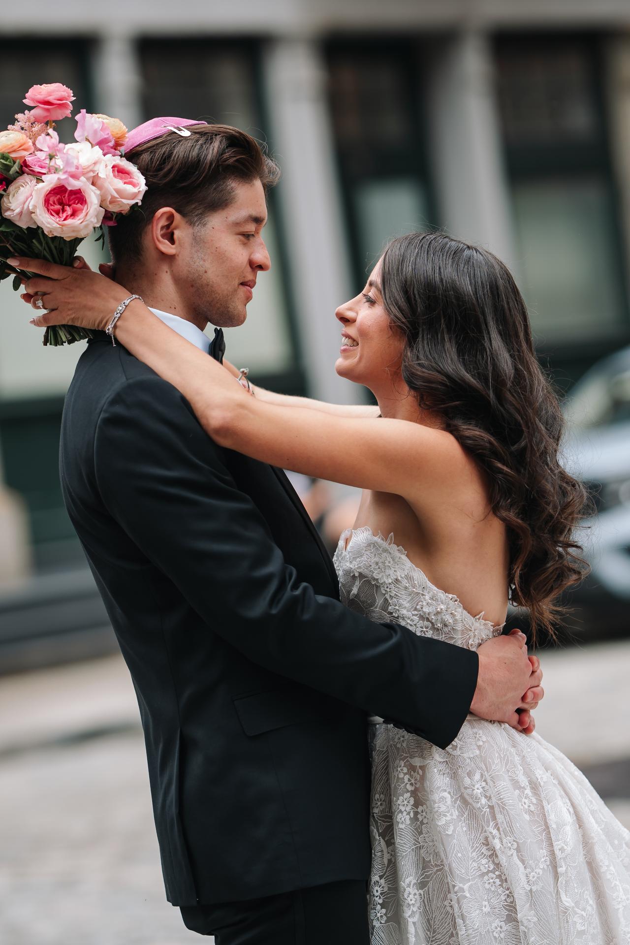 Bride and groom in an outdoor courtyard during badeken, embracing closely as the groom holds her hands and bouquet flowers fill the left side of the frame. Wedding photography at Tribeca Rooftop + 360°, New York, NY.