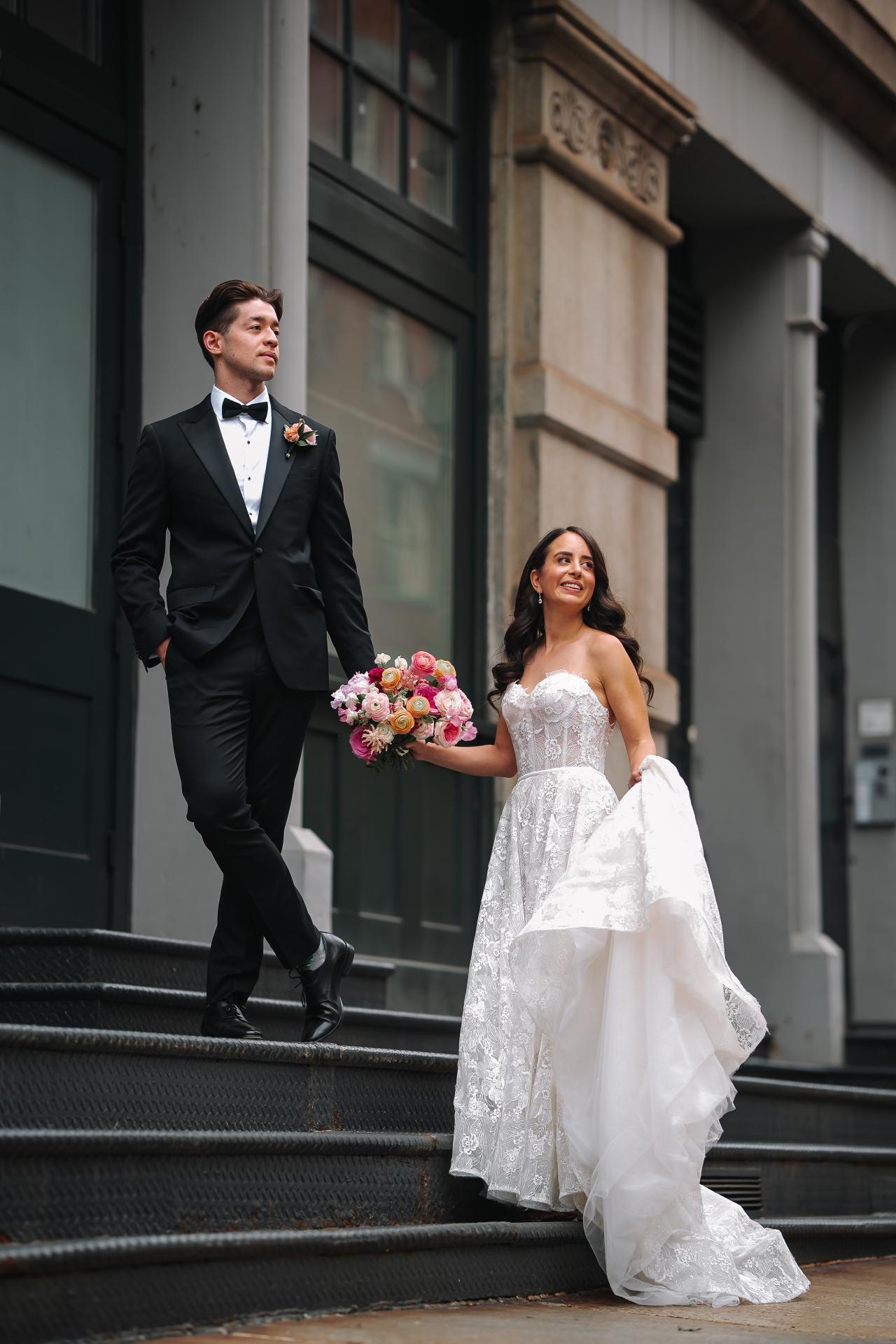 Bride in a white lace gown holding a colorful bouquet while the groom in a black tuxedo walks beside her on outdoor steps, framed by a stone-and-glass building façade. Wedding photography at Tribeca Rooftop + 360°, New York, NY.