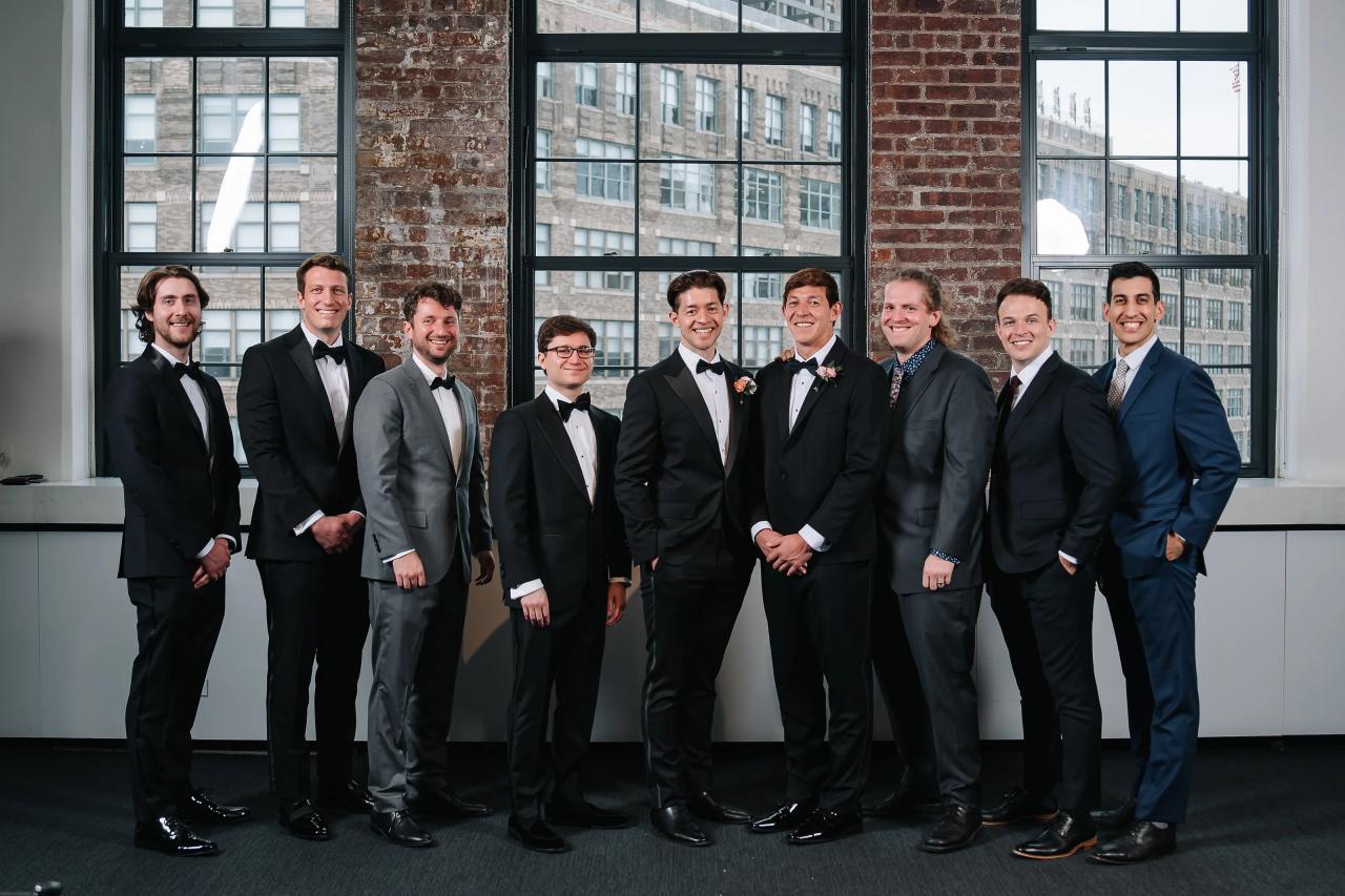 Posed group of men in formal suits and bow ties standing together in front of tall windows and exposed brick in an indoor venue. Wedding photography at Tribeca Rooftop + 360°, New York, NY.