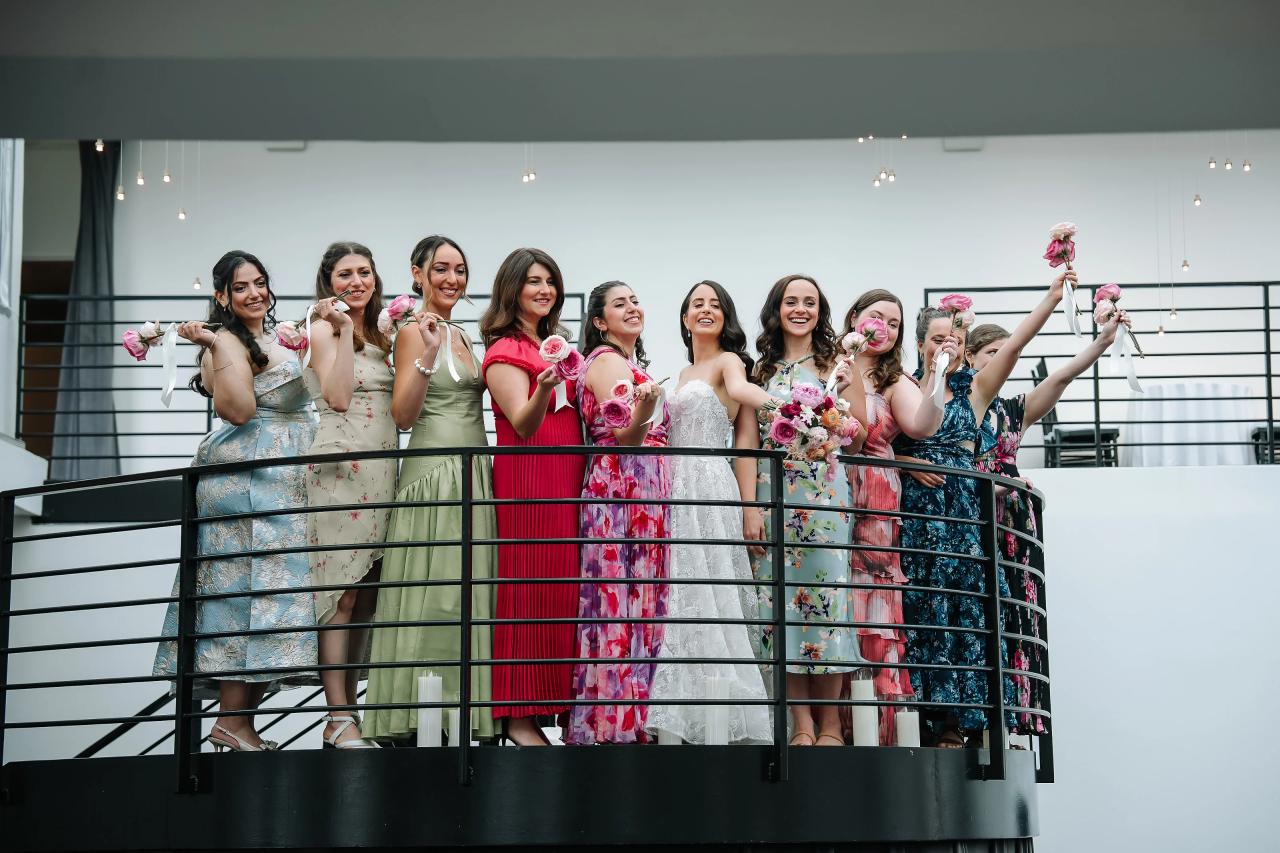 Group of women on an indoor balcony cheering and raising bouquets, captured mid-celebration with colorful matching outfits and warm overhead lights. Wedding photography at Tribeca Rooftop + 360°, New York, NY.