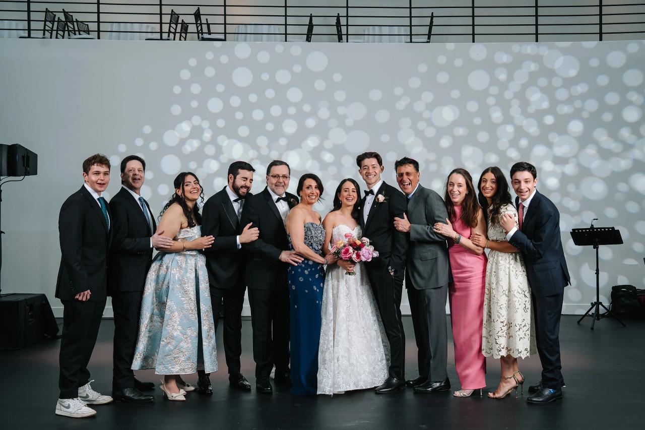Family group photo of a couple centered with friends and relatives smiling in front of a light-dot backdrop at an indoor reception. Wedding photography at Tribeca Rooftop + 360°, New York, NY.