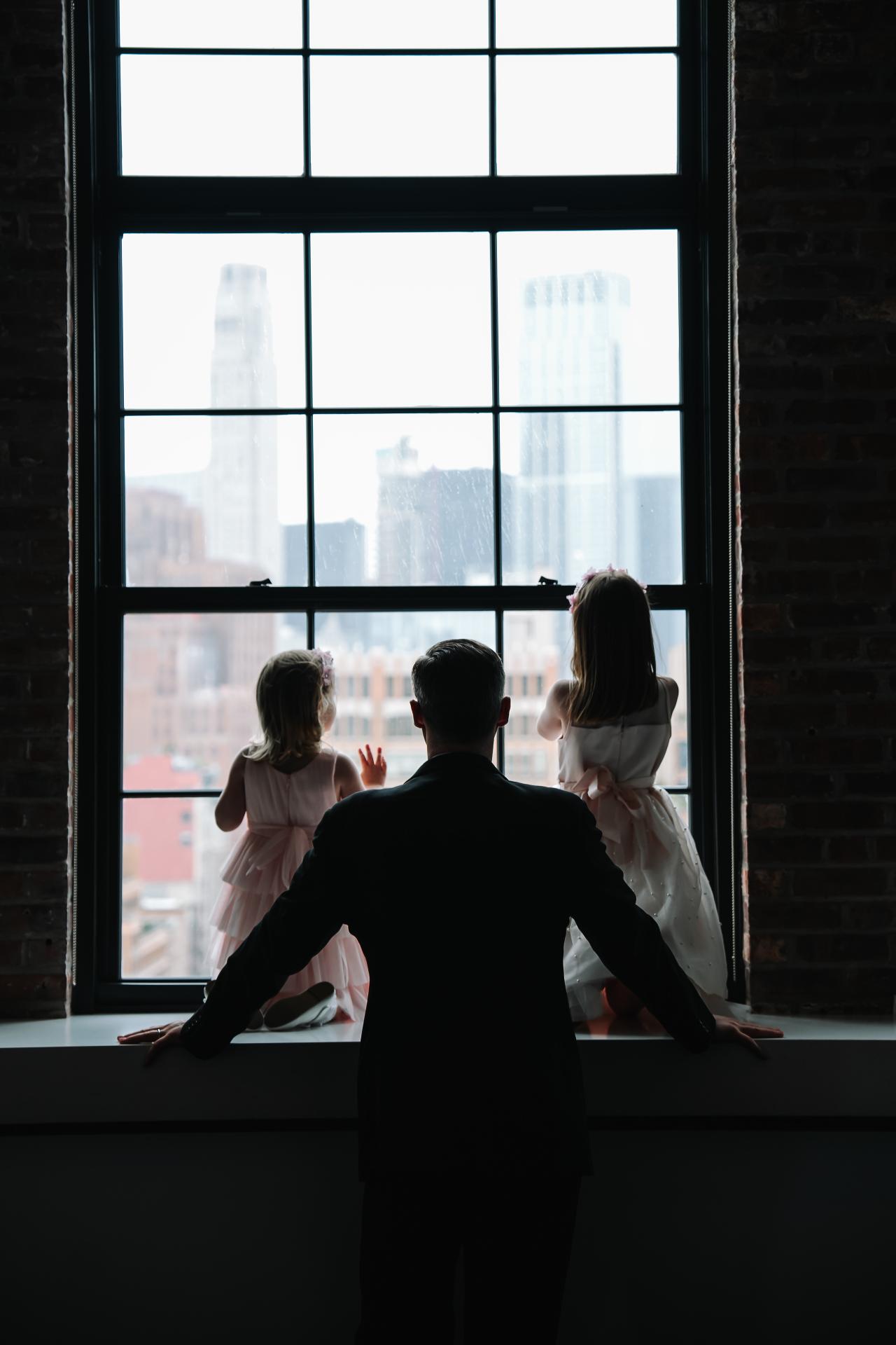 Silhouetted couple posed on a window ledge with raised hands, city skyline glowing in the background through a large grid window. Wedding photography at Tribeca Rooftop + 360°, New York, NY.