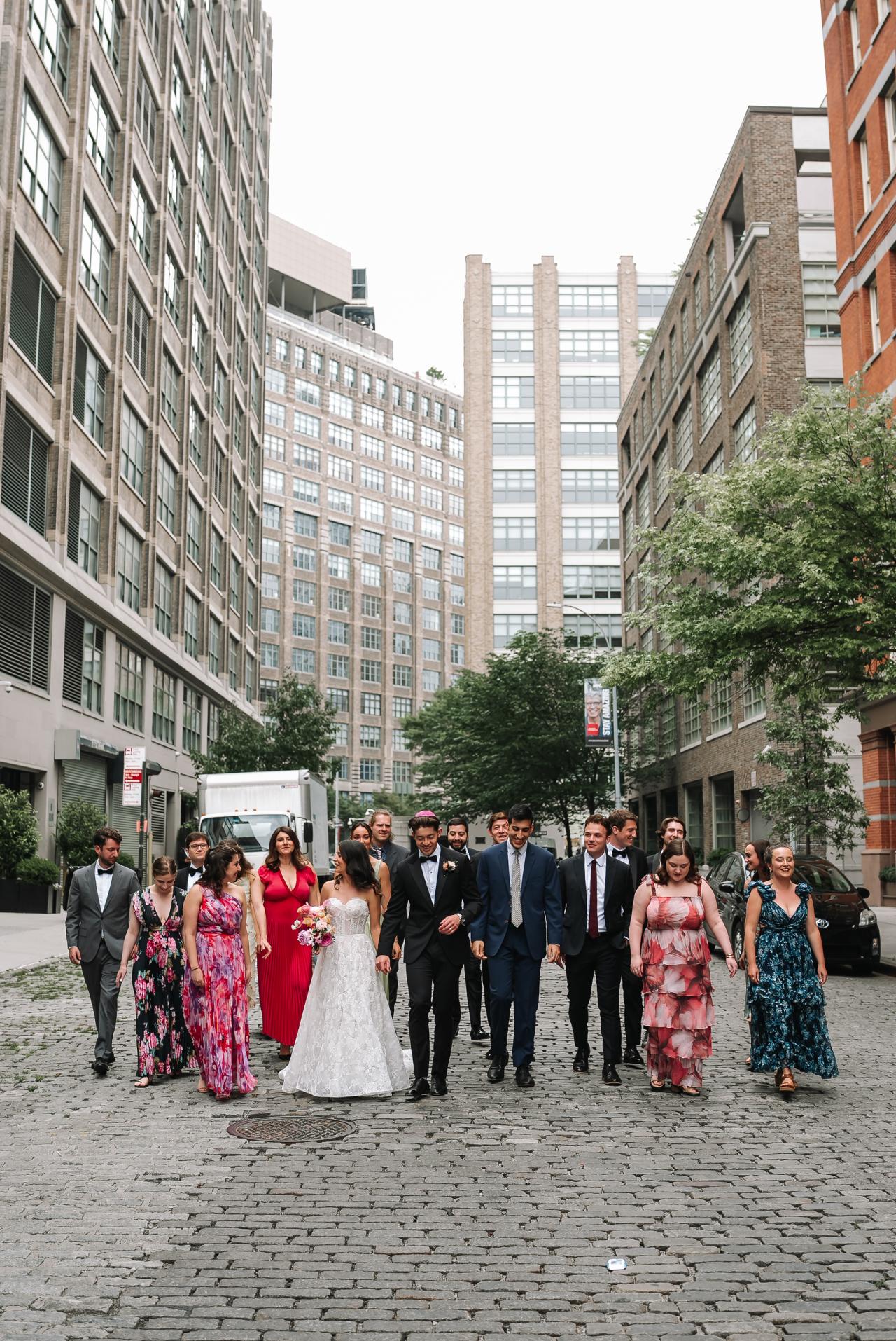 Wedding party walking together down a cobblestone street between tall buildings, bride and groom in the center of the line. Wedding photography at Tribeca Rooftop + 360°, New York, NY.