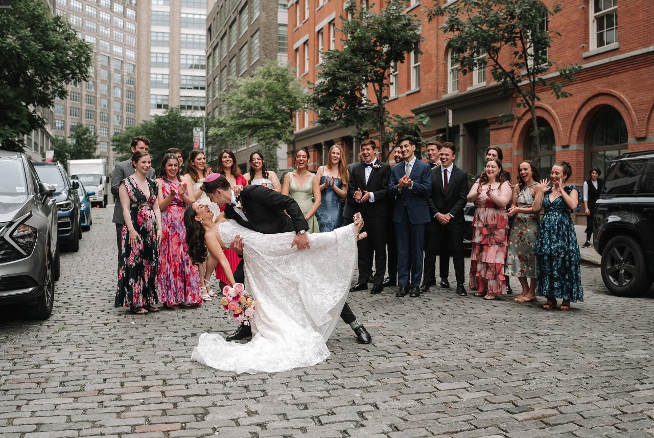 Wedding couple in a sweeping bridal gown captured mid-laugh while surrounded by clapping guests during an outdoor exit on a city street. Wedding photography at Tribeca Rooftop + 360°, New York, NY.