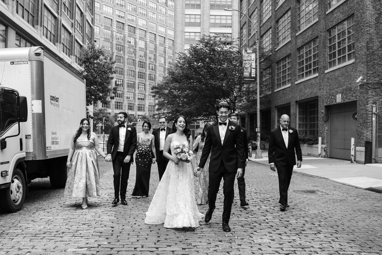 Black-and-white photo of a wedding couple walking down a cobblestone street outdoors, with formal guests following behind between tall brick buildings. Wedding photography at Tribeca Rooftop + 360°, New York, NY.