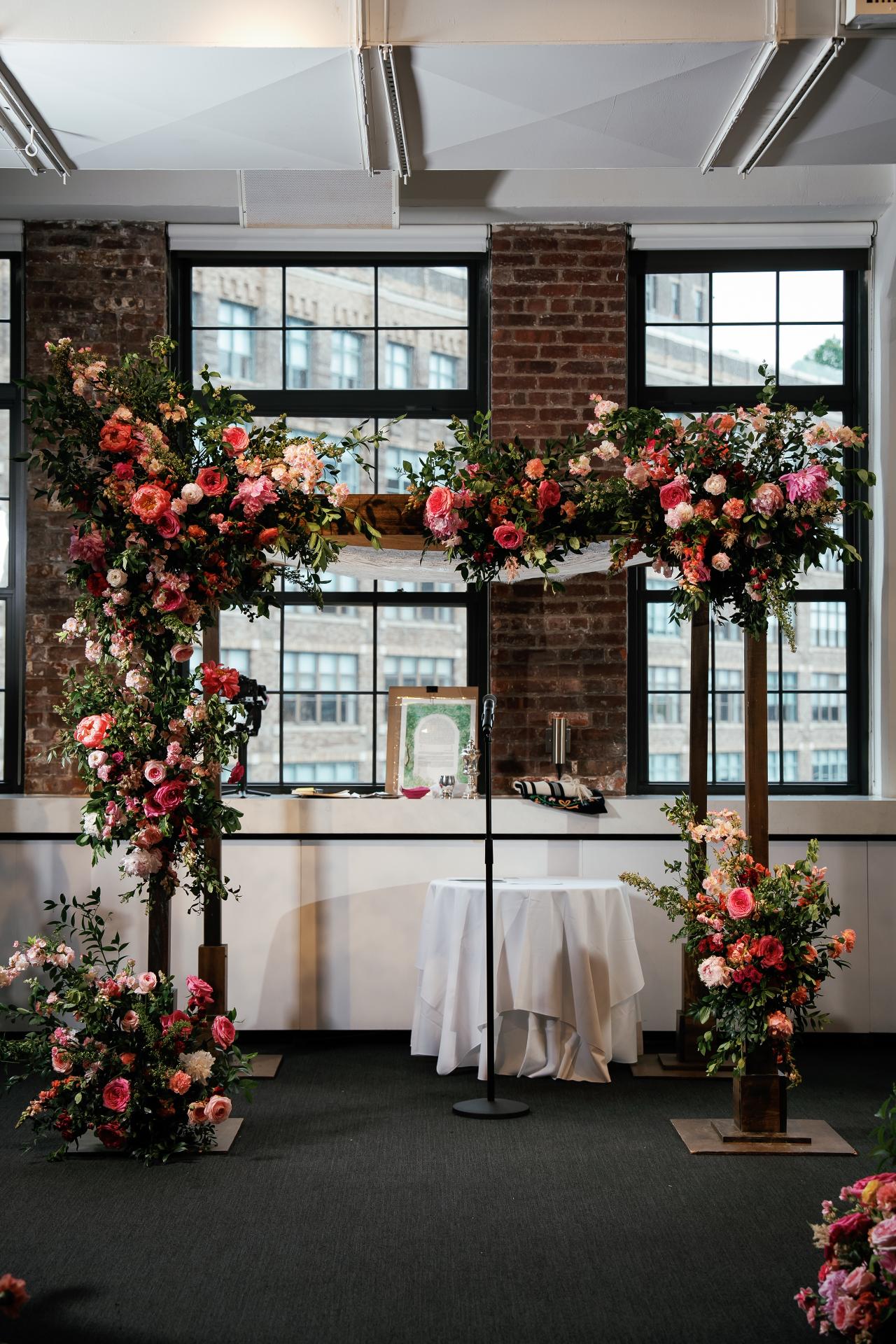 Floral chuppah ceremony setup with red and pink rose arrangements and greenery, framed by tall stands in a bright indoor room with brick and large windows. Wedding photography at Tribeca Rooftop + 360°, New York, NY.