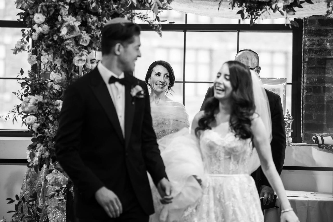 Black-and-white candid of a bride smiling while seated with groom standing nearby, flowers and window light framing the scene, with a softly blurred guest in the foreground. Wedding photography at Tribeca Rooftop + 360°, New York, NY.