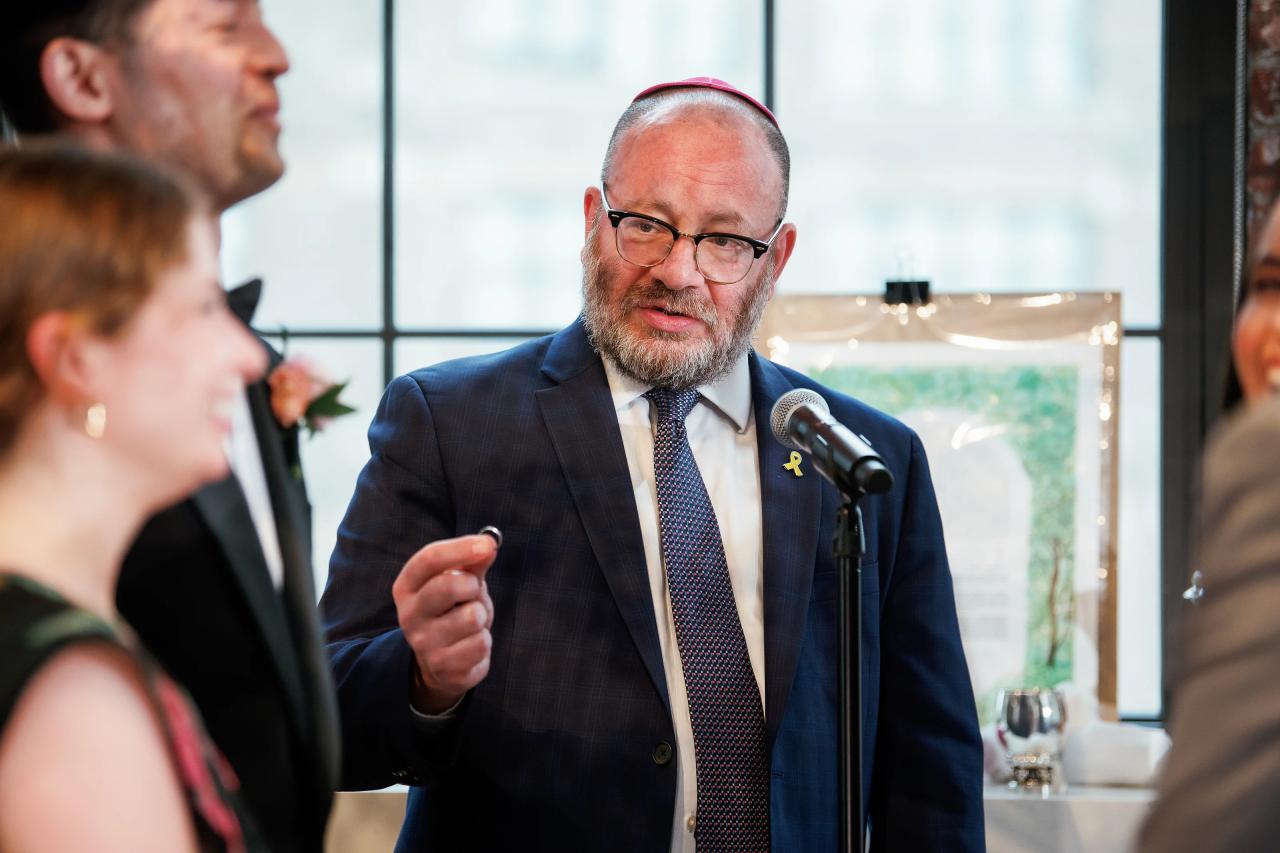 Man in a suit speaking into a microphone at an event, with audience members blurred in the foreground. Wedding photography at Tribeca Rooftop + 360°, New York, NY.