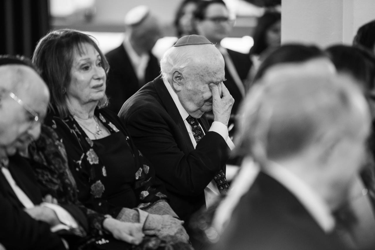 Black-and-white candid of an older man sitting at a celebration with his hand to his face, an older woman beside him looking on, with guests blurred in the background. Wedding photography at Tribeca Rooftop + 360°, New York, NY.