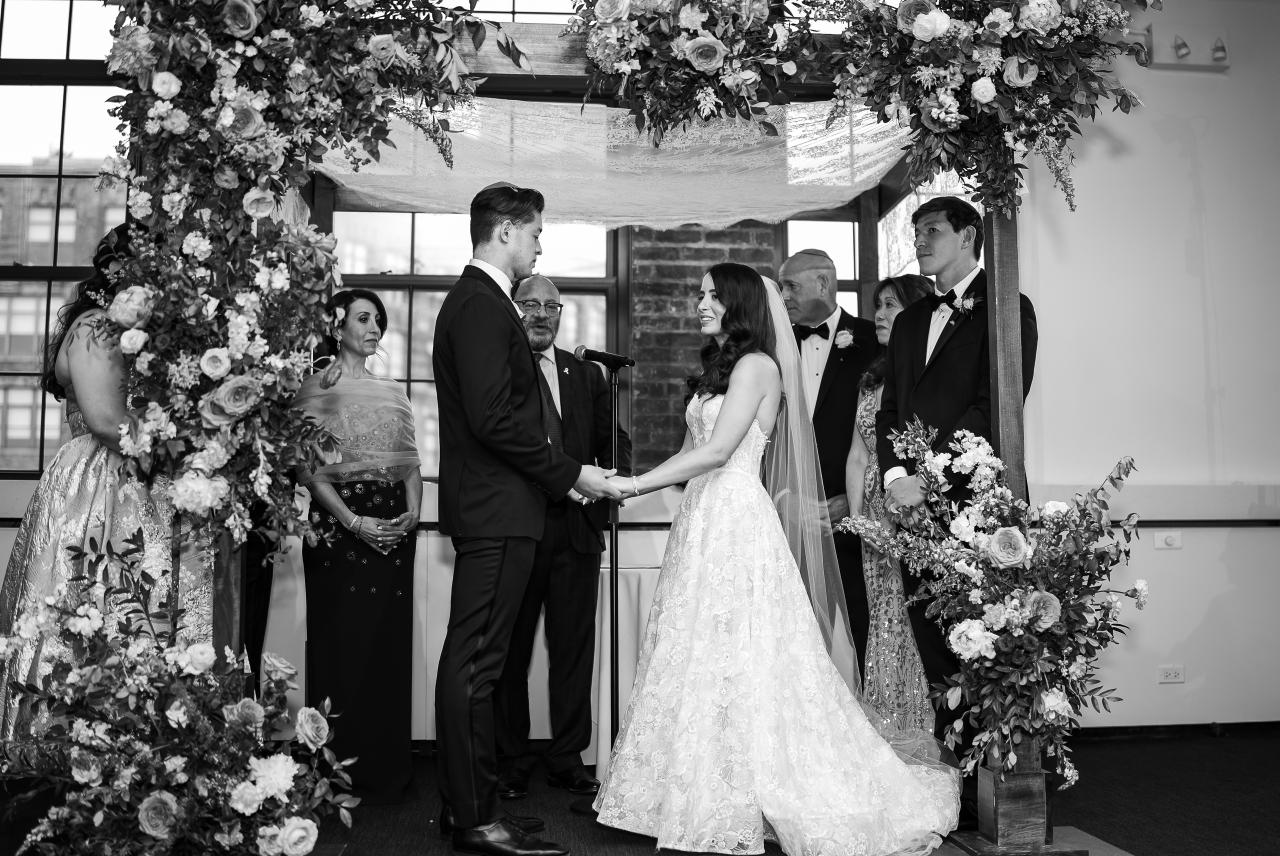 Black-and-white ceremony photo of a couple holding hands beneath an ornate floral canopy, with wedding guests in formal attire standing in the background. Wedding photography at Tribeca Rooftop + 360°, New York, NY.