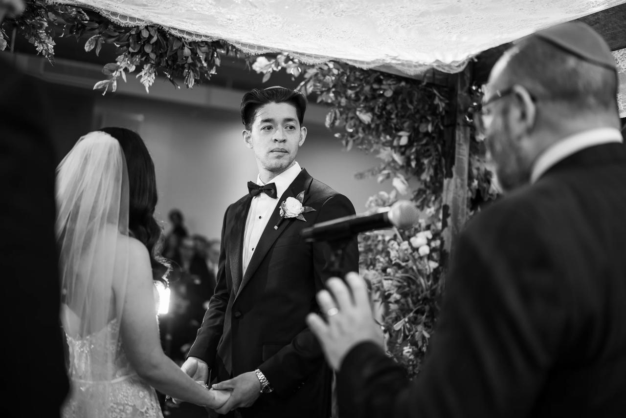 Black-and-white wedding photograph of a groom in a tuxedo and bow tie standing with hands clasped as the bride in a veil looks toward him, with an officiant’s gesture in the foreground under floral décor. Wedding photography at Tribeca Rooftop + 360°, New York, NY.