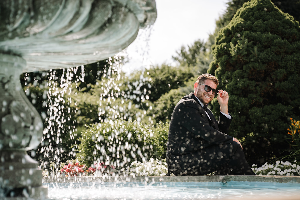 Groom laughing and adjusting his ear/hat while sitting poolside, with water splashing and floral decor in the background.