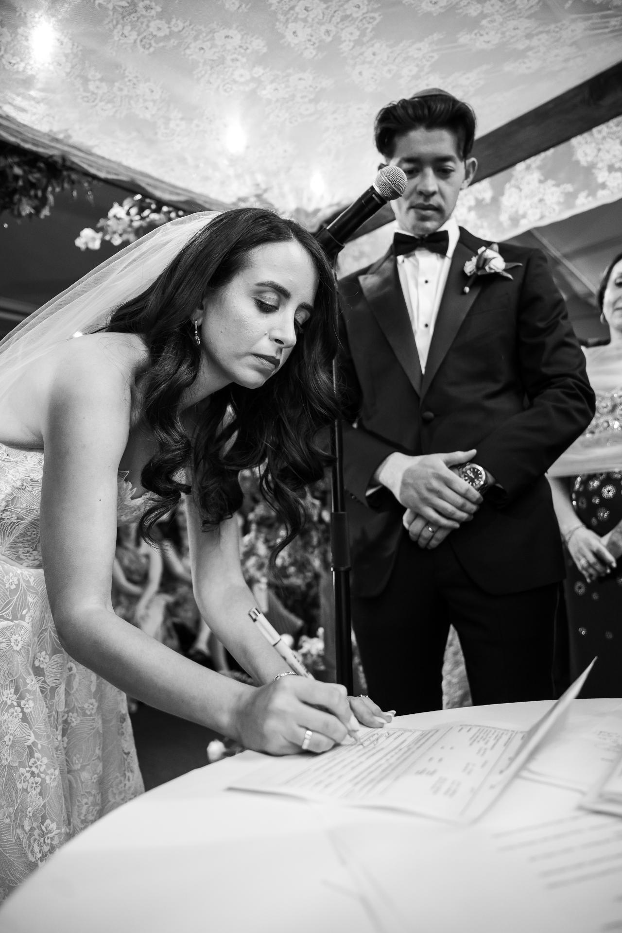 Black-and-white ceremony moment: bride signing a document with the groom standing behind her under a canopy, microphone in front, guests blurred in the background. Wedding photography at Tribeca Rooftop + 360°, New York, NY.