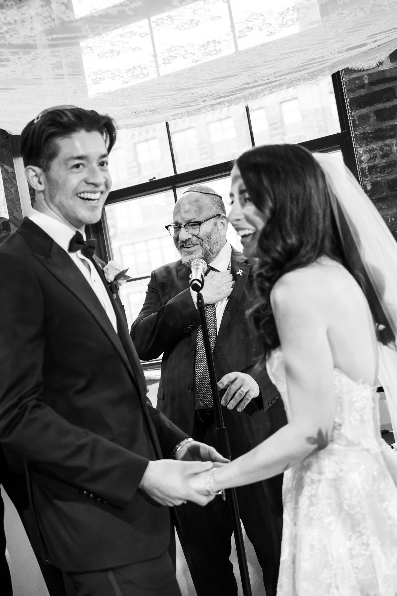 Black-and-white close ceremony moment: a man speaks into a microphone while the bride and groom stand facing him under a canopy, sharing a smiling, intimate exchange. Wedding photography at Tribeca Rooftop + 360°, New York, NY.