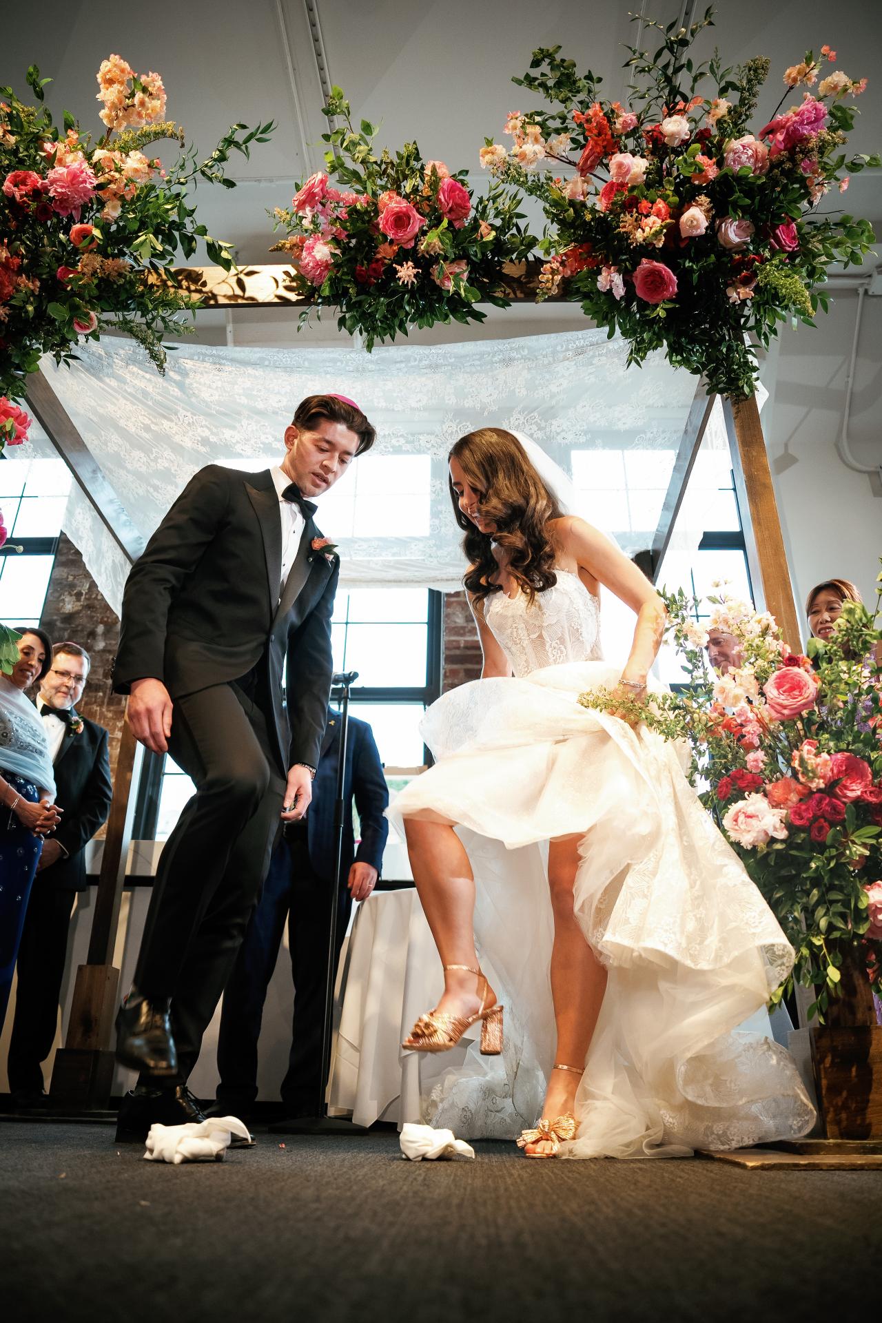 Bride and groom at the chuppah, bride adjusting her dress while the groom stands beside her, framed by floral arrangements in a bright indoor ceremony space. Wedding photography at Tribeca Rooftop + 360°, New York, NY.