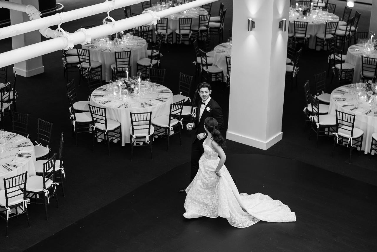 Black-and-white wedding reception finale: bride in a white gown and groom in a tuxedo walking past reception tables in an open dance area. Wedding photography at Tribeca Rooftop + 360°, New York, NY.