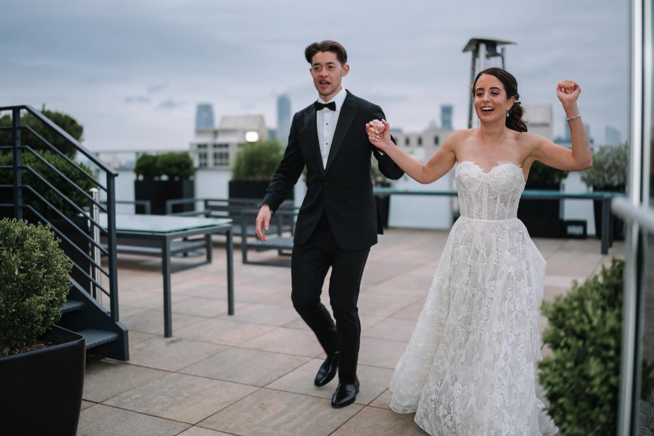 Wedding couple in formal attire celebrating and dancing outdoors, holding hands with joyful expressions against a city skyline backdrop. Wedding photography at Tribeca Rooftop + 360°, New York, NY.
