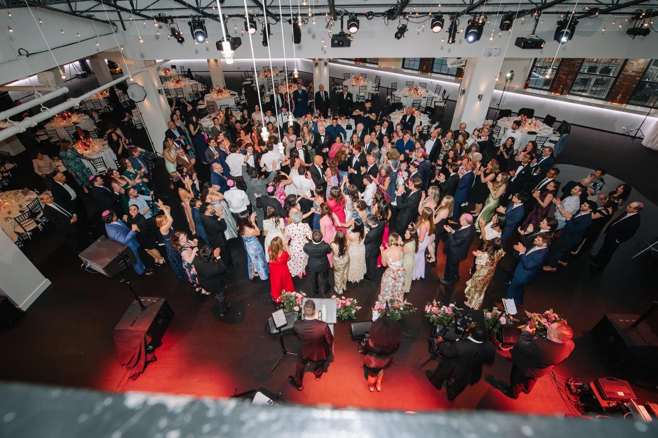 Overhead wide shot of a large crowd celebrating at an indoor event, with guests applauding around the central couple on a red carpet. Wedding photography at Tribeca Rooftop + 360°, New York, NY.