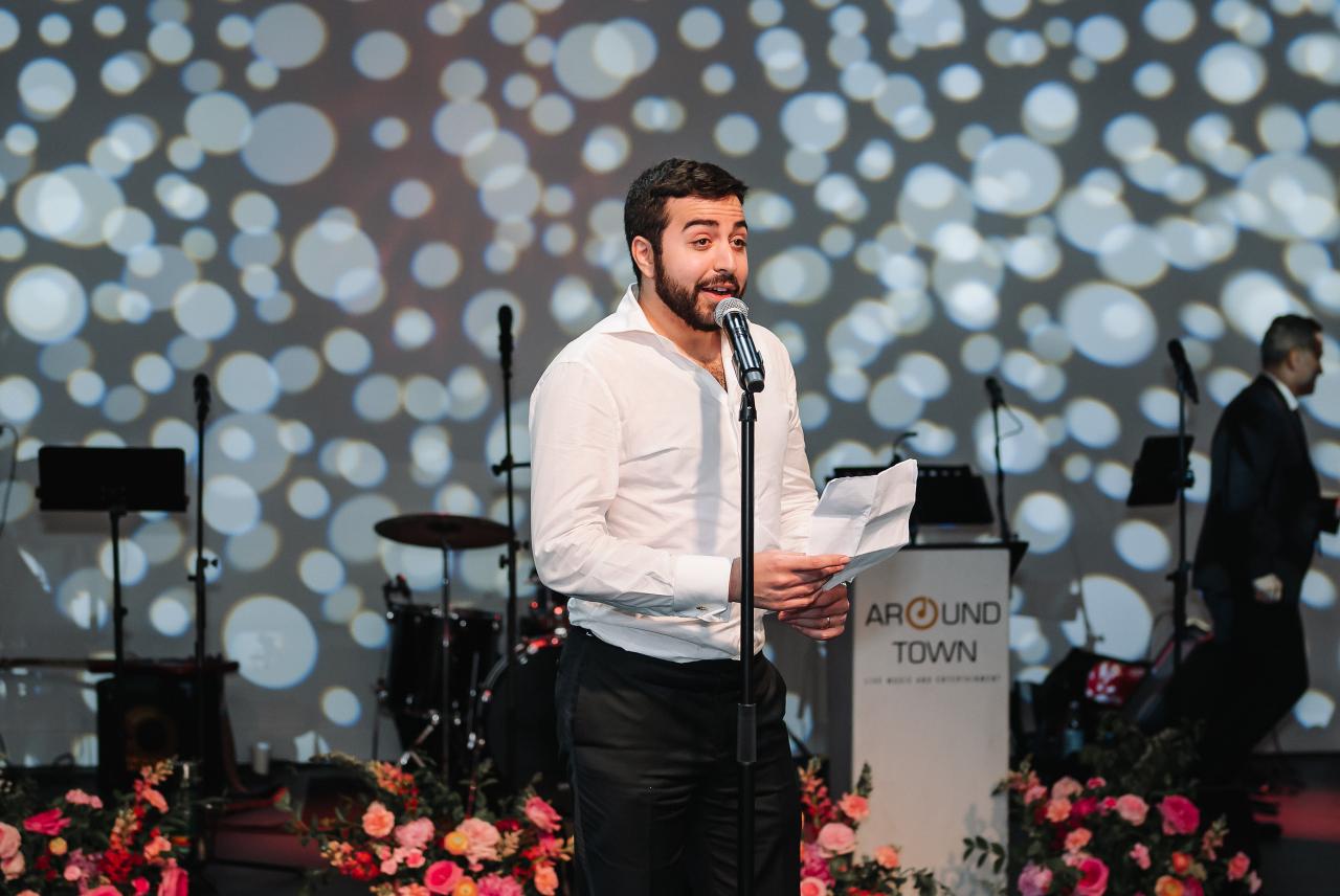 Man in a white shirt speaking into a microphone on a wedding event stage, holding notes, with a wall of glowing circular lights behind him and floral arrangements below. Wedding photography at Tribeca Rooftop + 360°, New York, NY.