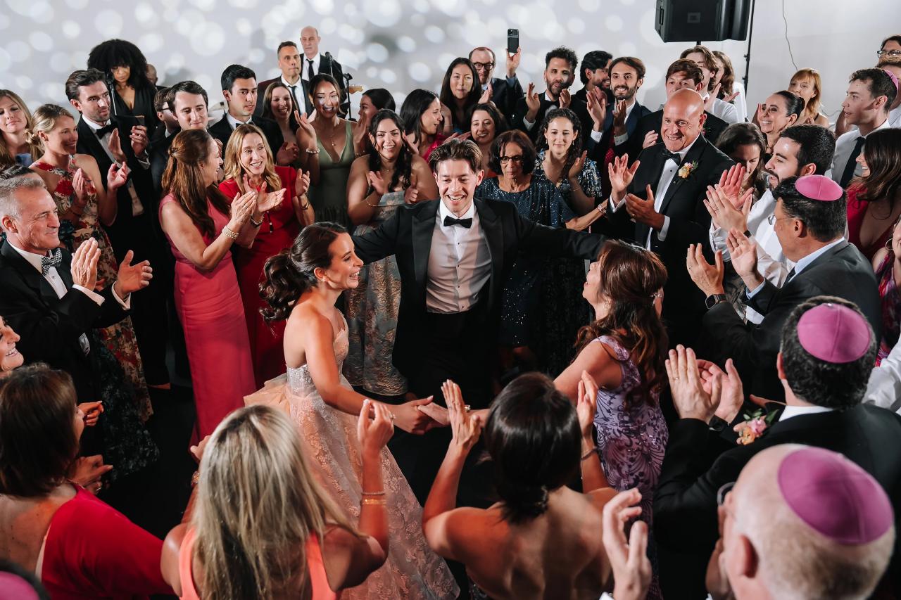 Bride and groom smiling in the center as guests surround them in a joyful clap and cheers at a wedding celebration. Wedding photography at Tribeca Rooftop + 360°, New York, NY.