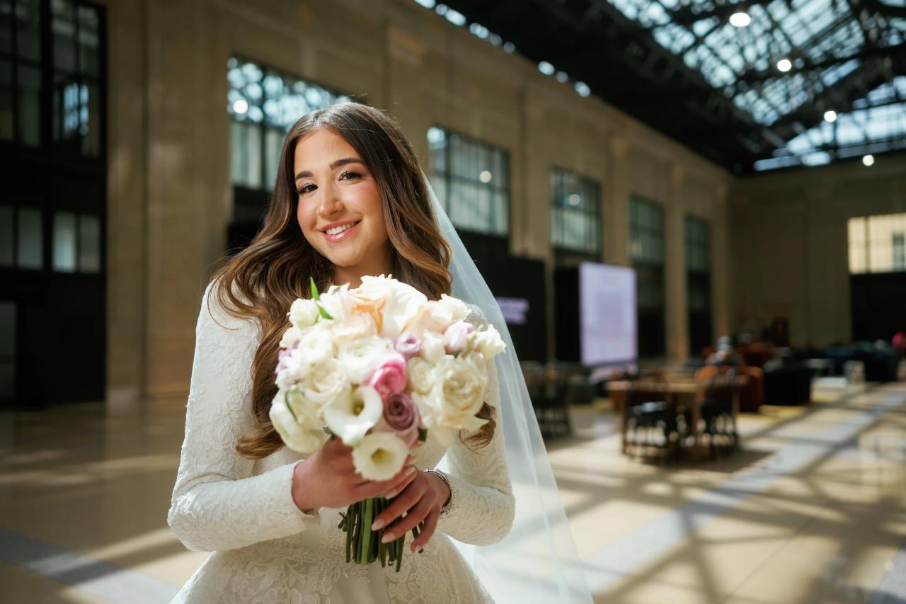 Smiling bride holding a bouquet of white and blush-pink flowers in bright indoor light, with veil framing her face.