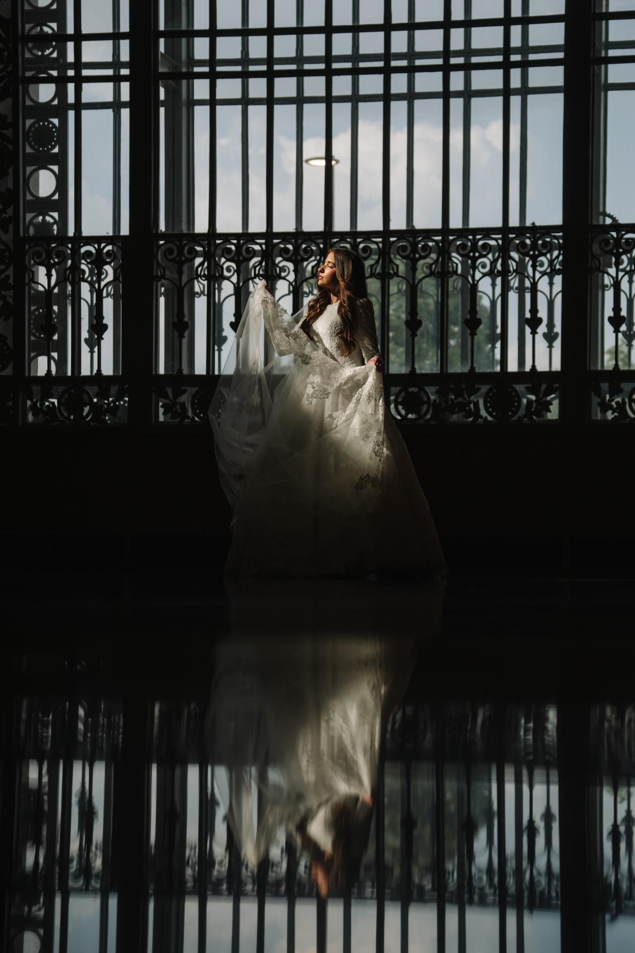 Bride in a flowing gown and veil silhouetted against strong backlight behind ornate window ironwork, with a clear reflection on a polished floor.