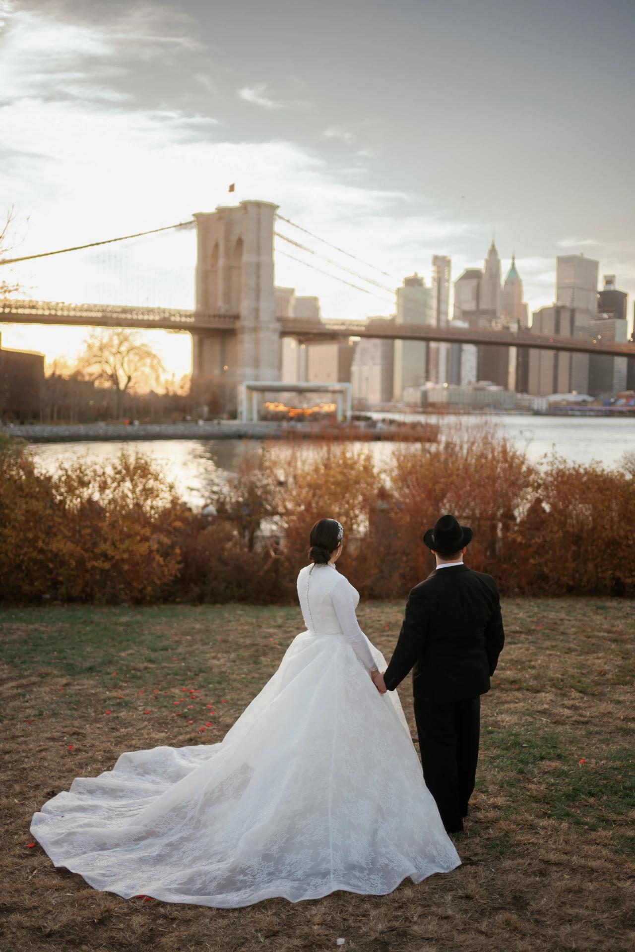 Bride and groom walking hand in hand outdoors at sunset with the Brooklyn Bridge and city skyline in the background; the bride’s veil and gown train trail behind her.
