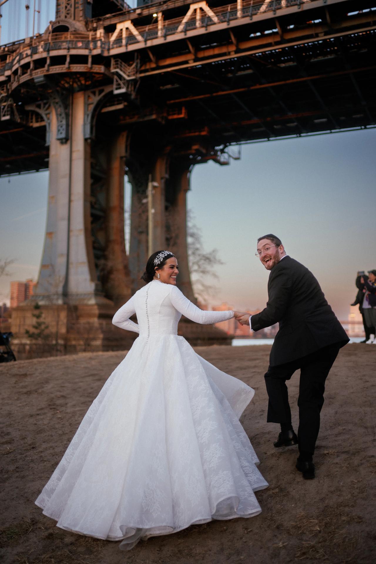 Outdoor wedding photo of a bride in a white gown and a groom in a dark suit holding hands and smiling while walking, with a grand arched building in the background.