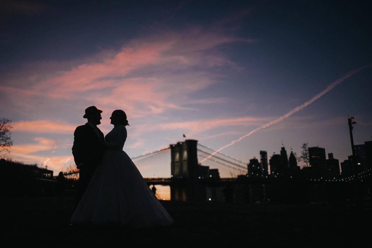 Silhouetted bride and groom holding each other at dusk with a lit city skyline and bridge in the background.
