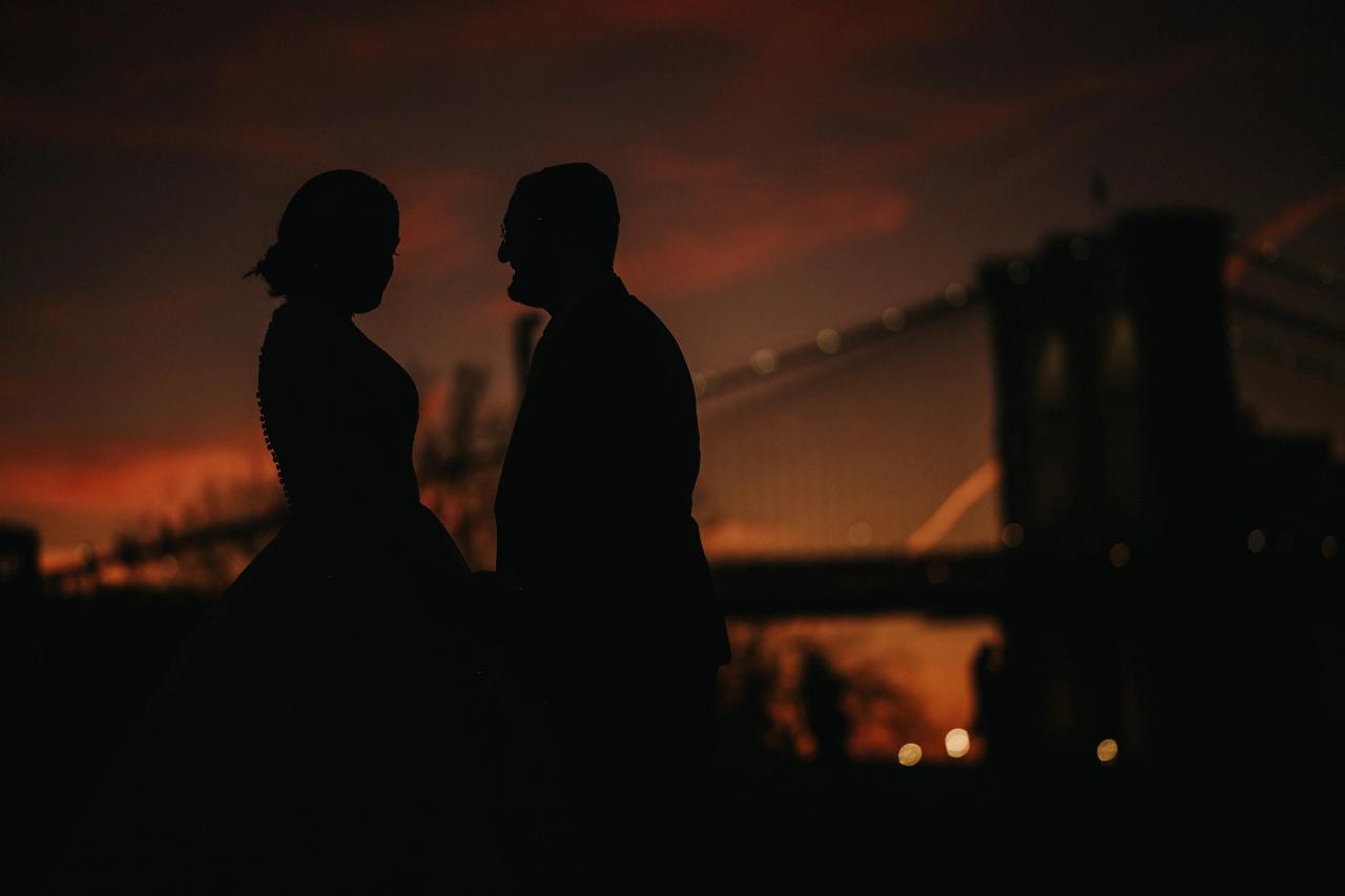 Silhouetted bride and groom standing close together in profile during a nighttime reception, with warm amber lights and bokeh in the background.