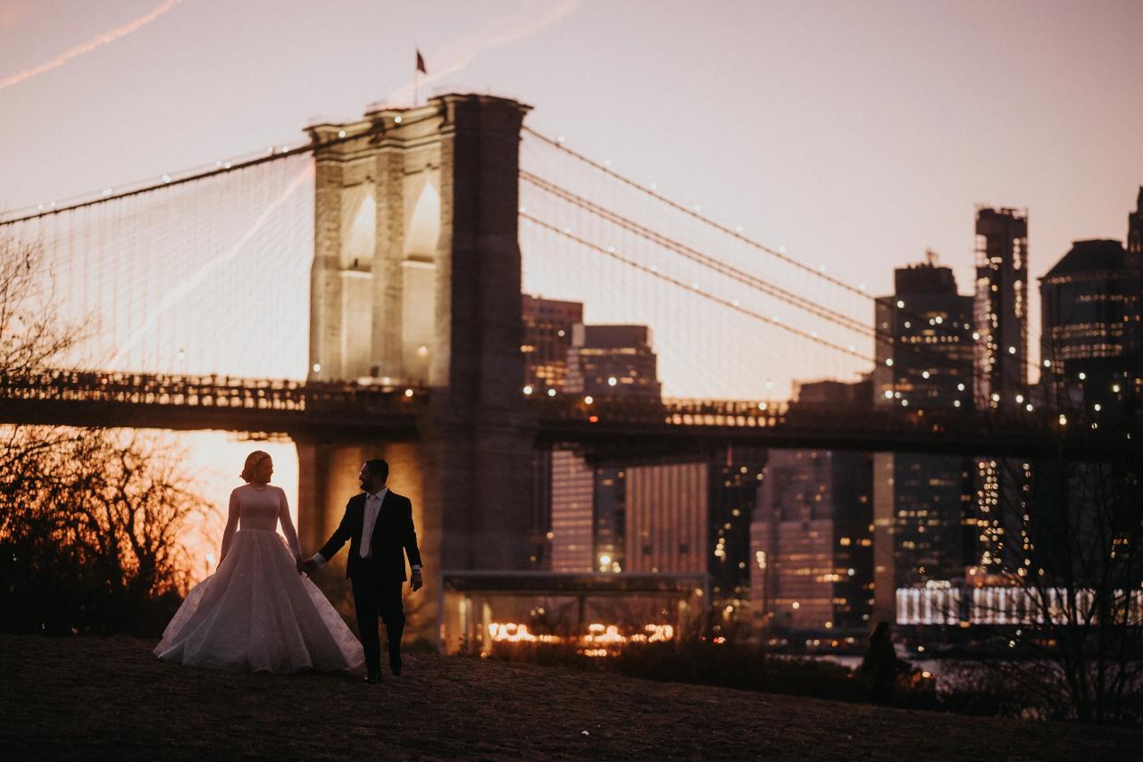 Bride and groom walking hand-in-hand outdoors at golden hour with a lit suspension bridge and skyline in the background.