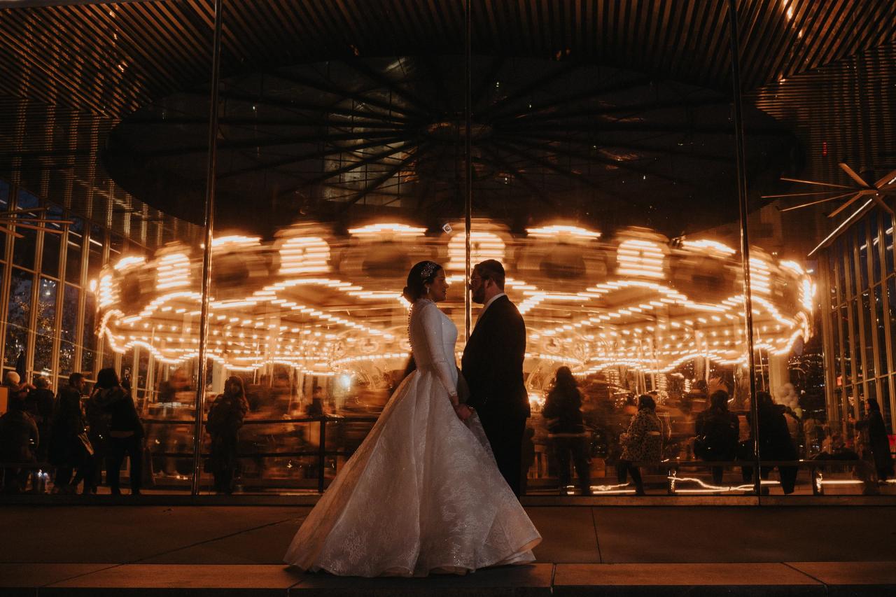 Framed by warm LED lights and light trails, a bride and groom walk together holding hands during a lively indoor celebration at night.